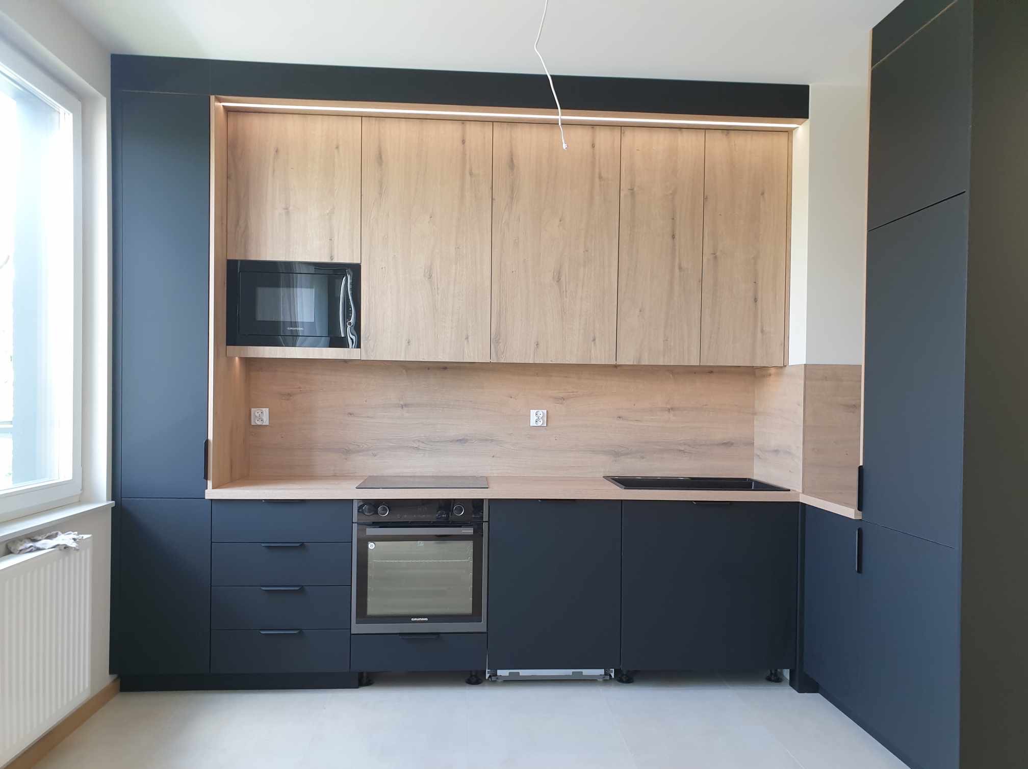Nowoczesna kuchnia z ciemnogranatowymi szafkami dolnymi i górnymi wykończonymi drewnopodobną okleiną, oświetlenie LED podszafkowe, mikrofalówka w zabudowie.