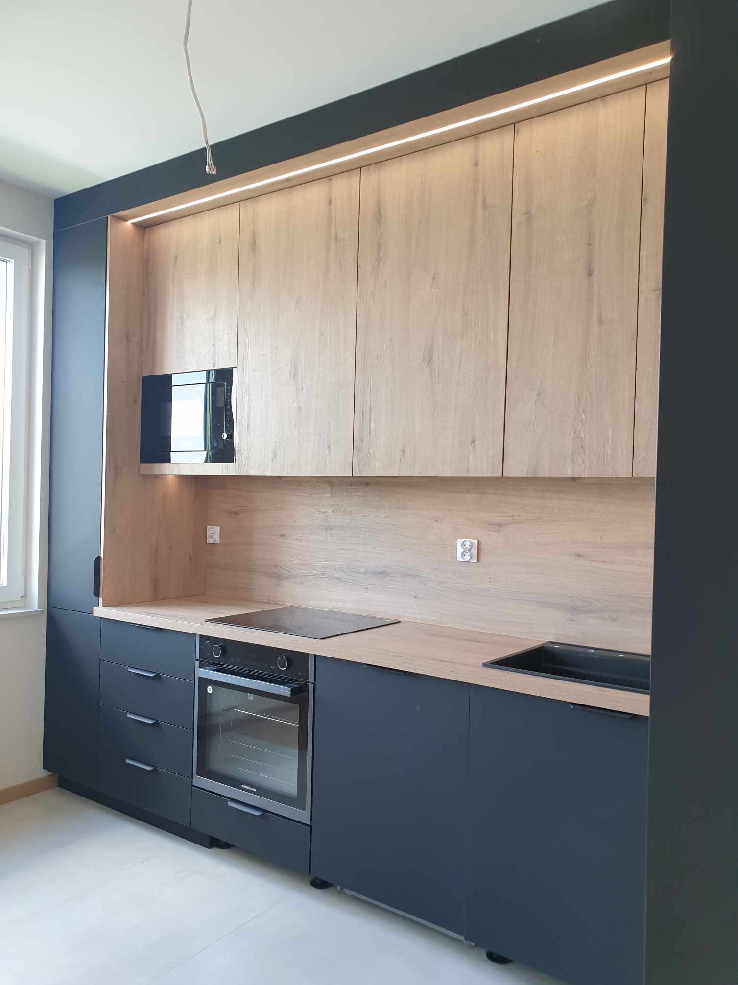 Nowoczesna kuchnia z drewnianymi frontami szafek górnych i ciemnogranatowymi dolnymi, oświetlenie LED podszafkowe, czarny zlew i płyta indukcyjna, wbudowana mikrofalówka.