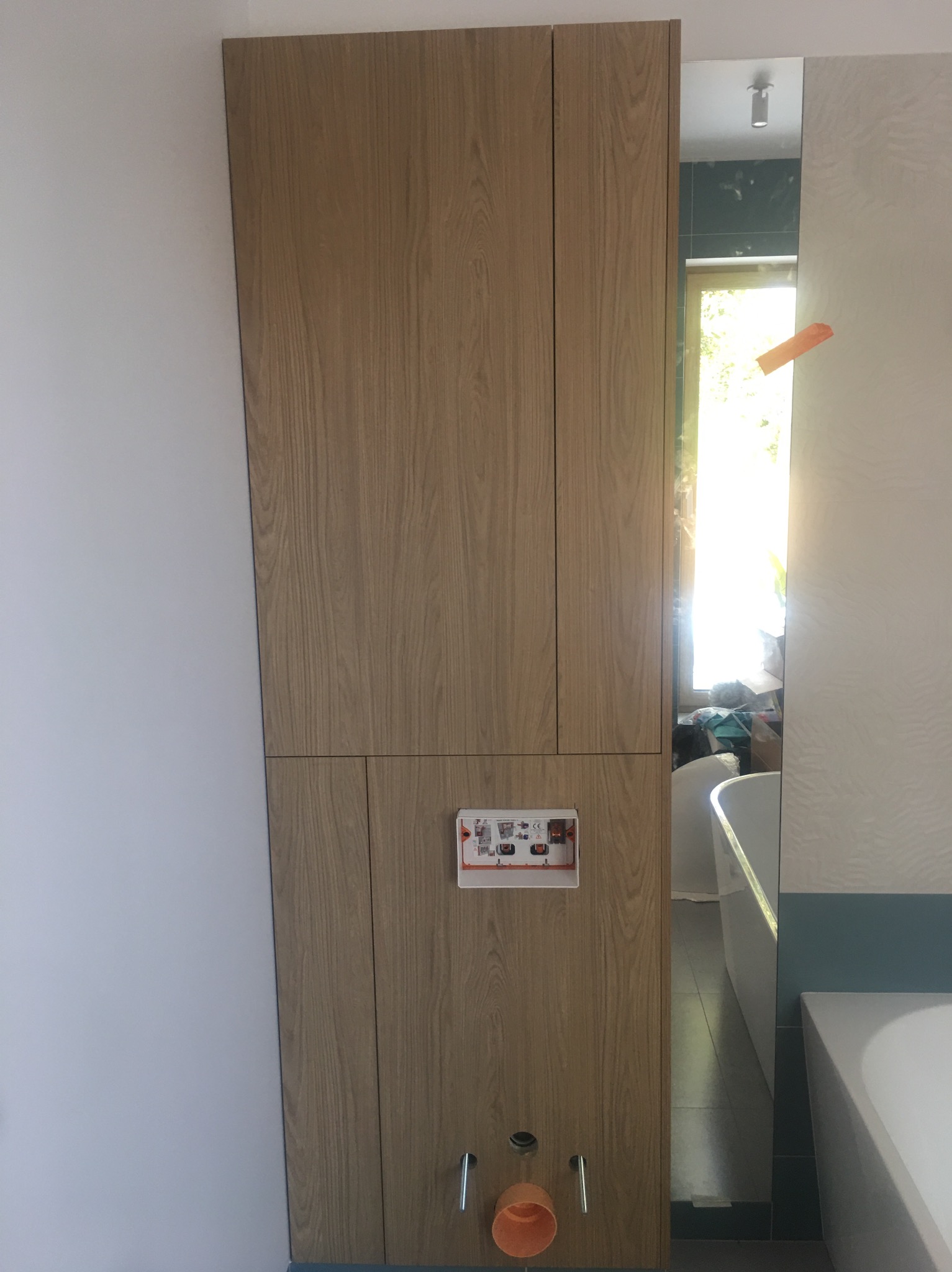 Drewniana zabudowa stelaża WC z przyciskiem spłukującym i elementami instalacji, widoczna wanna w tle.