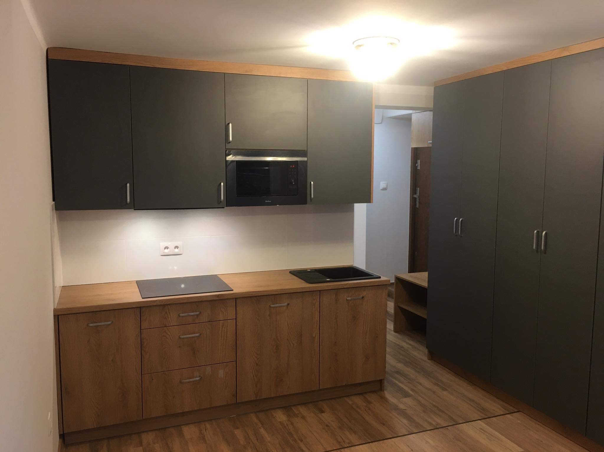 Nowoczesna kuchnia: szare szafki górne z drewnianą listwą, drewniane szafki dolne, czarny zlew i płyta indukcyjna, oświetlenie podszafkowe, duża szafa w zabudowie po prawej stronie.