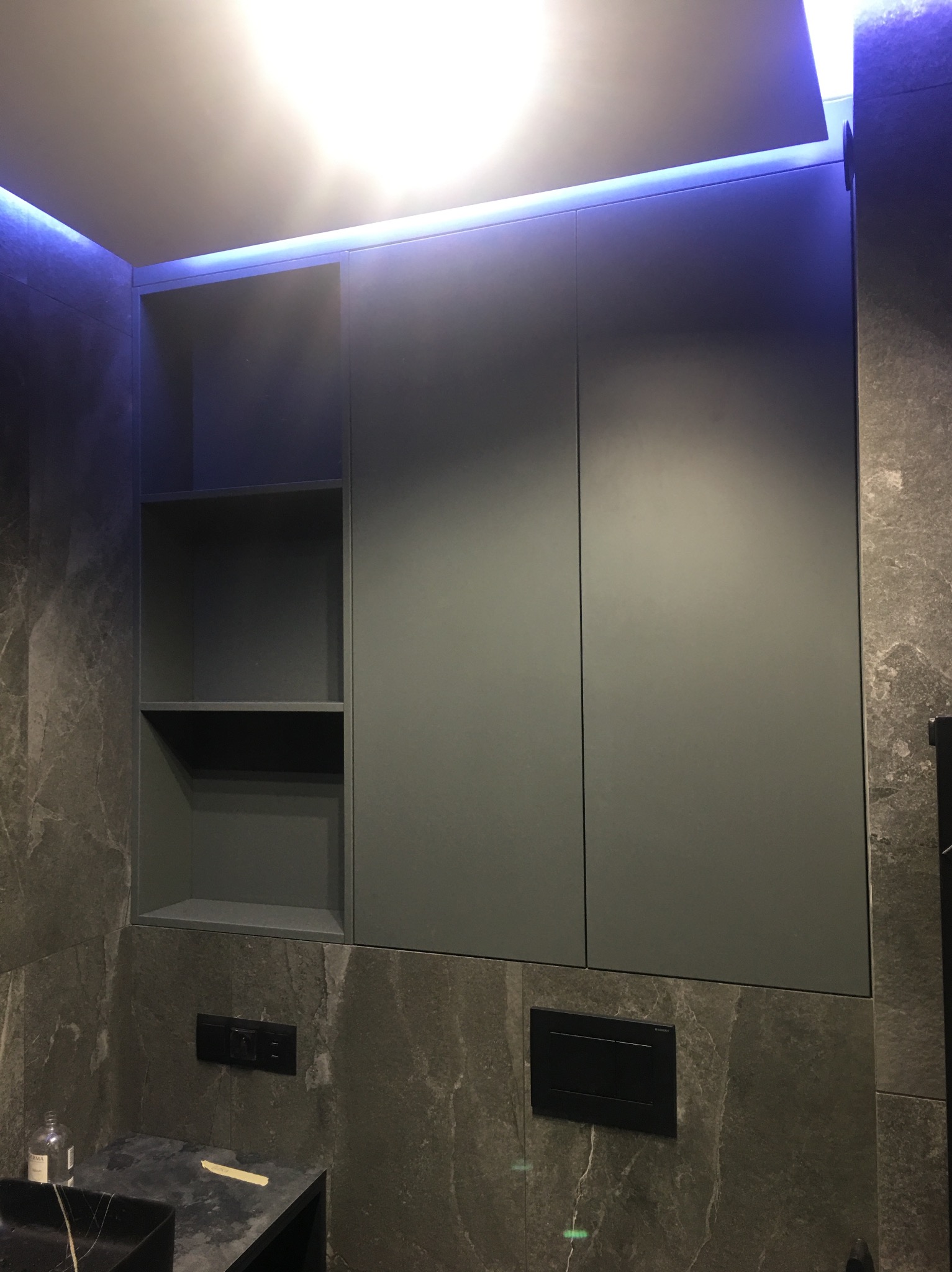 Szare szafki łazienkowe na wymiar z oświetleniem LED, marmurowe ściany, czarny blat umywalki.