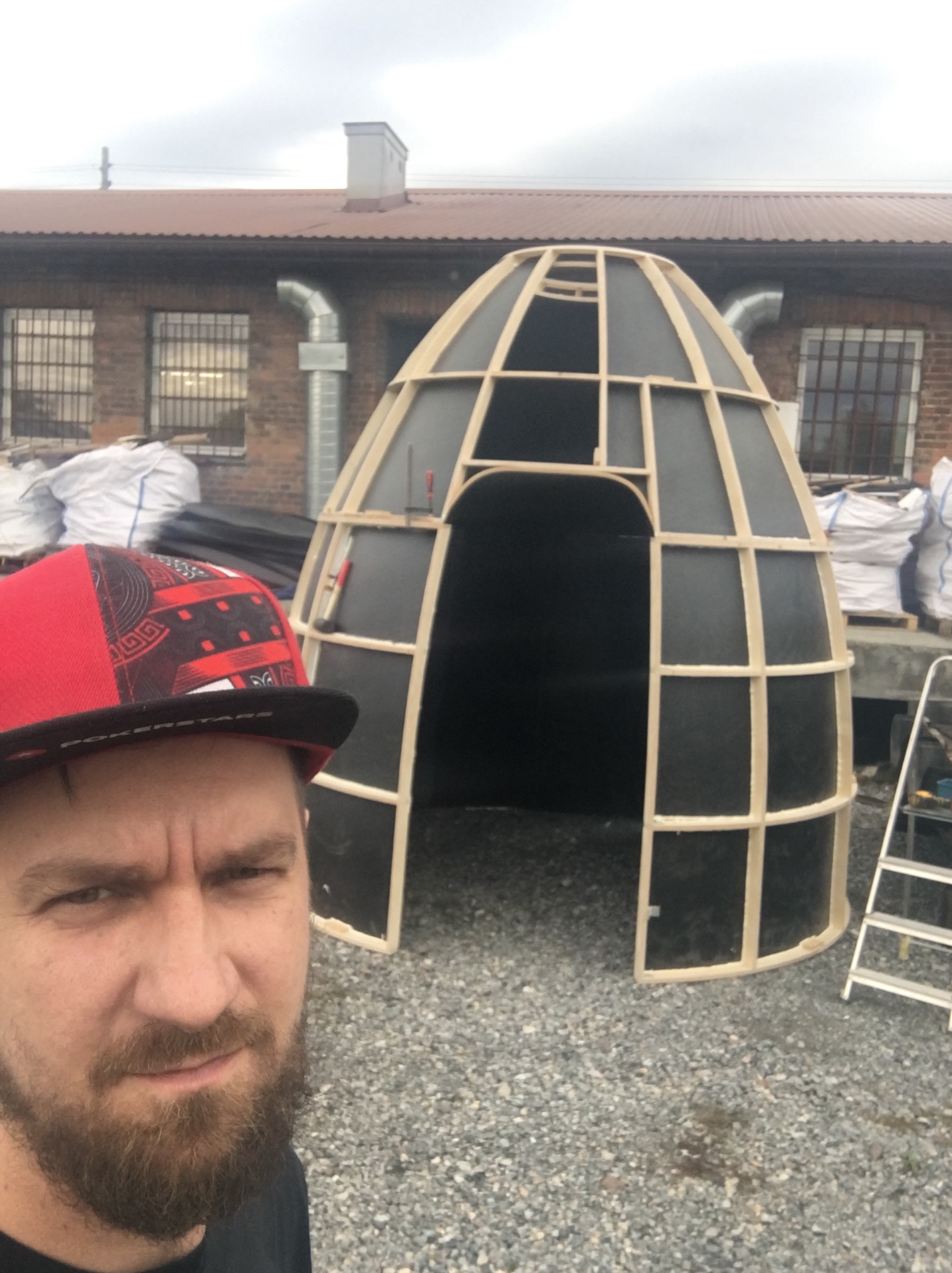 Mężczyzna w czapce robi selfie na tle szkieletu konstrukcji w kształcie igloo z drewnianych belek i czarnych paneli, w tle budynek z cegły i worki z materiałami budowlanymi.