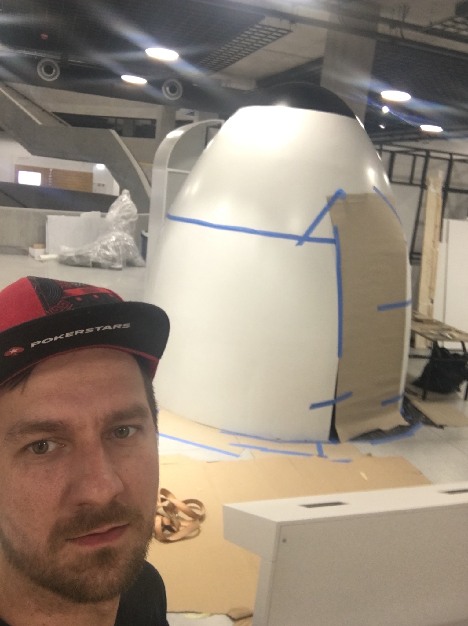 Selfie mężczyzny w czapce PokerStars na tle dużej, białej, jajowatej konstrukcji z niebieską taśmą malarską i kartonem, prawdopodobnie w trakcie budowy w przestronnym, industrialnym wnętrzu.