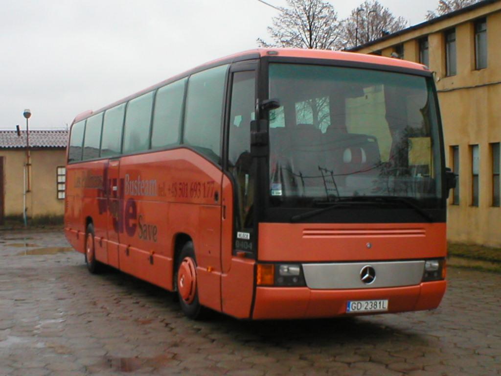 Pomarańczowy autobus Mercedes z napisem 'Busteam' i numerem telefonu, zaparkowany na brukowanym placu przed budynkiem w pochmurny dzień.