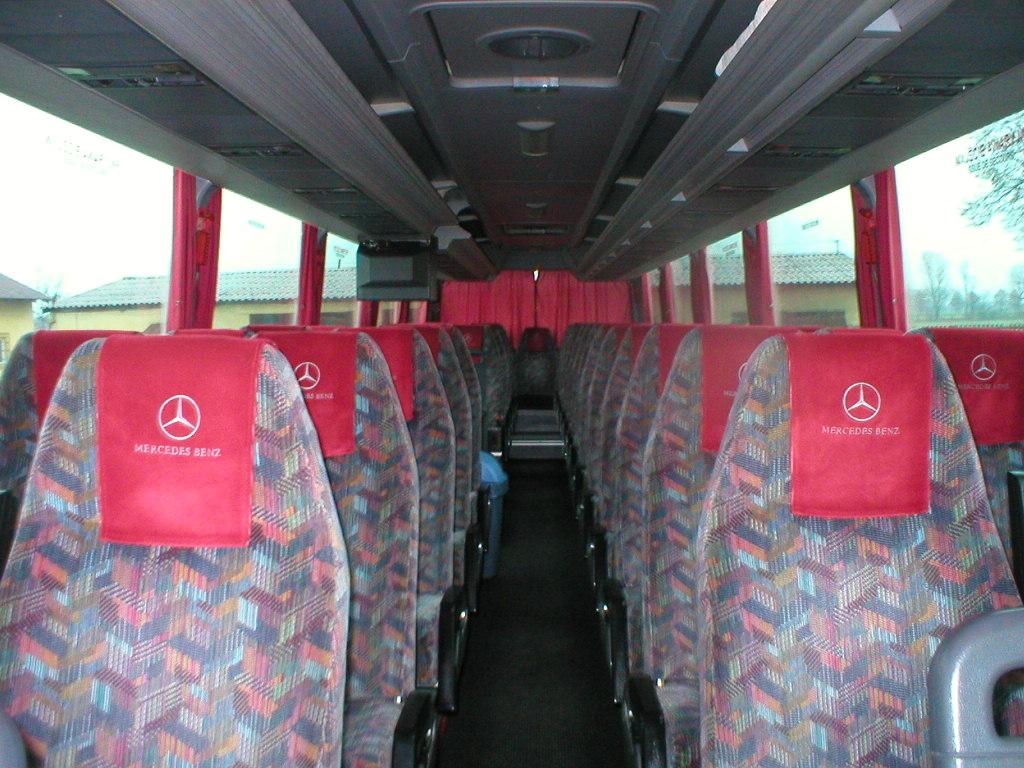 Wnętrze autobusu Mercedes-Benz z rzędami foteli obitych wzorzystą tkaniną w odcieniach czerwieni, niebieskiego i szarości, z czerwonymi zagłówkami z logo Mercedes-Benz.