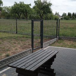 BRAMEX Przemysław Tabaka - Ciemna ławka z drewnianych bali na kostce brukowej, na tle ogrodzenia panelowego z bramką i zieleni. Pochmurne niebo.