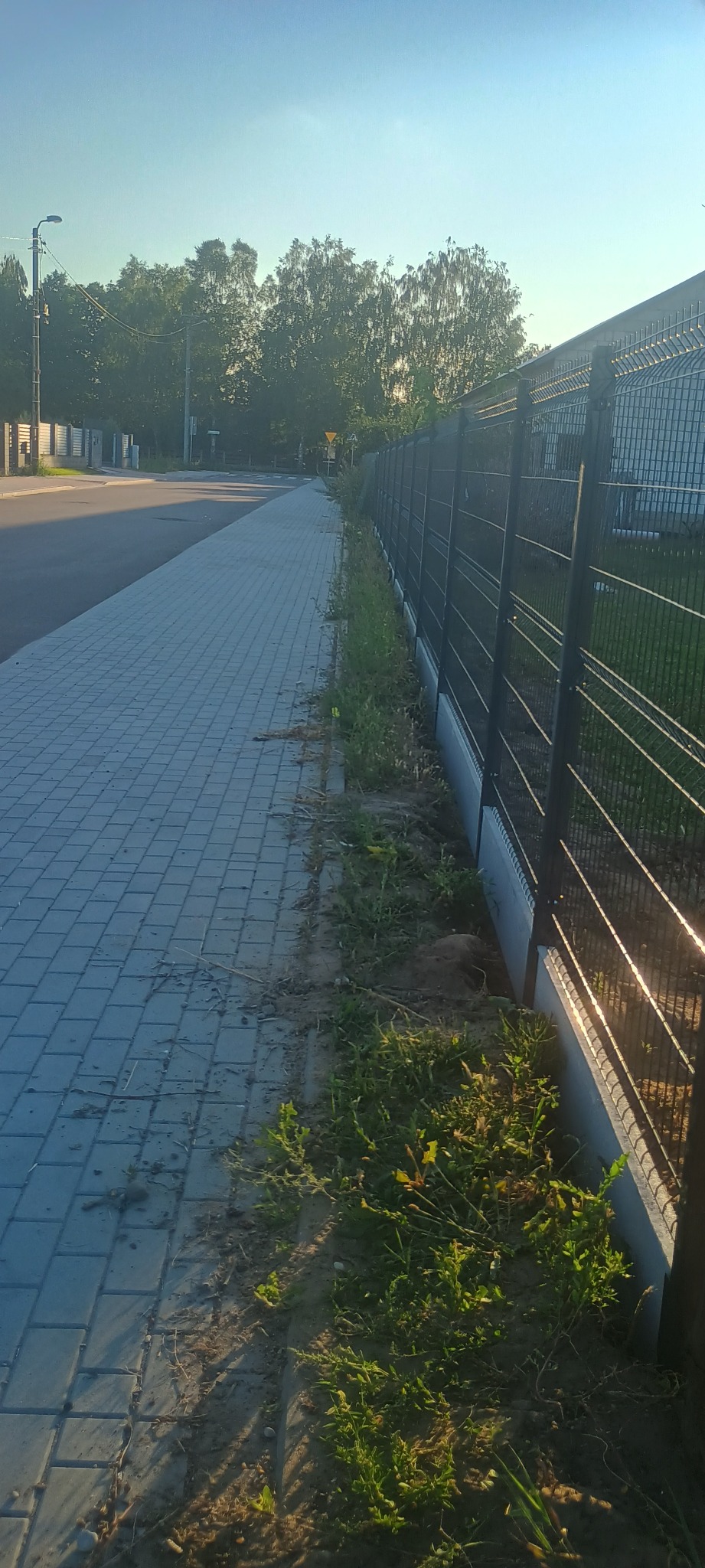 Długi, ciemny, metalowy płot panelowy z betonową podmurówką, widoczny z poziomu chodnika z kostki brukowej w słoneczny dzień.