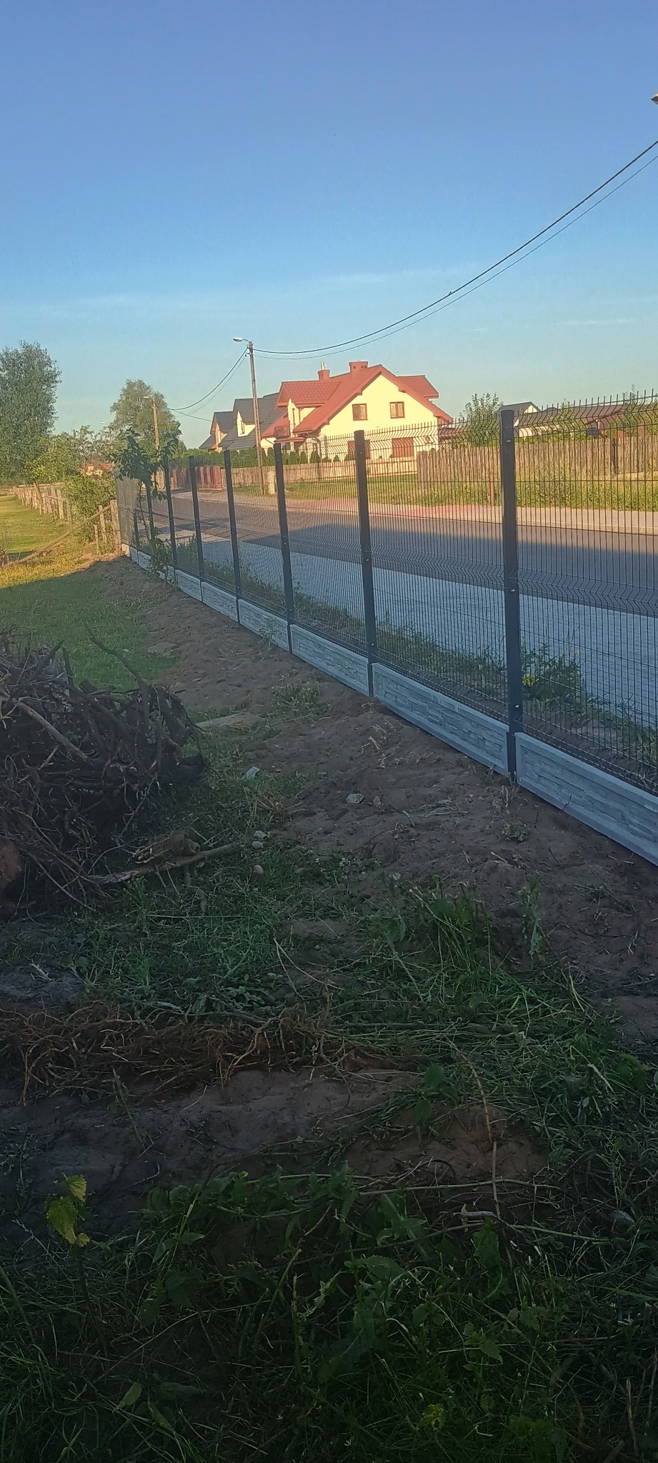 Nowoczesne ogrodzenie panelowe z podmurówką imitującą kamień, widok na ulicę i domy w tle.