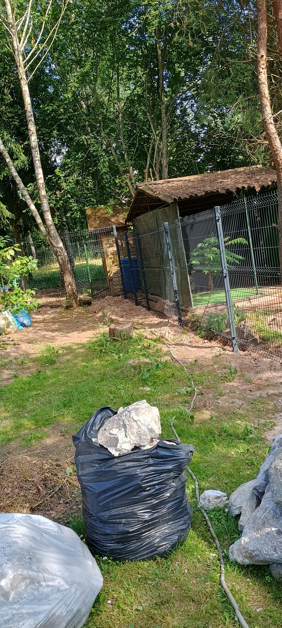 Fragment ogrodzenia z siatki w Kadzidle, z widocznym workiem na śmieci i kamieniami na pierwszym planie, teren zarośnięty trawą i drzewami.