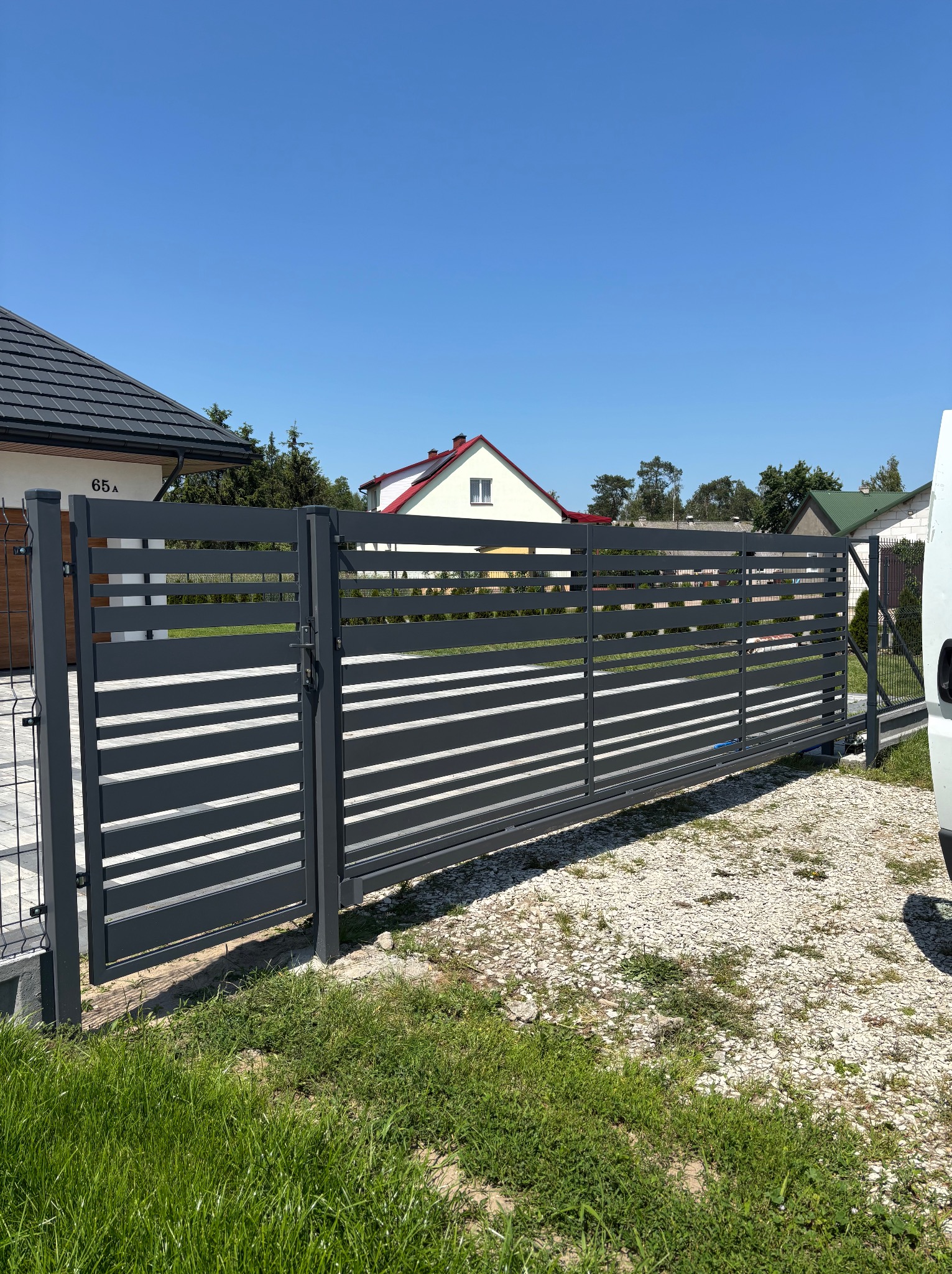 Nowoczesna, przesuwna brama w kolorze antracytowym z poziomymi panelami, zamontowana przed domem z numerem 65A, na tle błękitnego nieba.