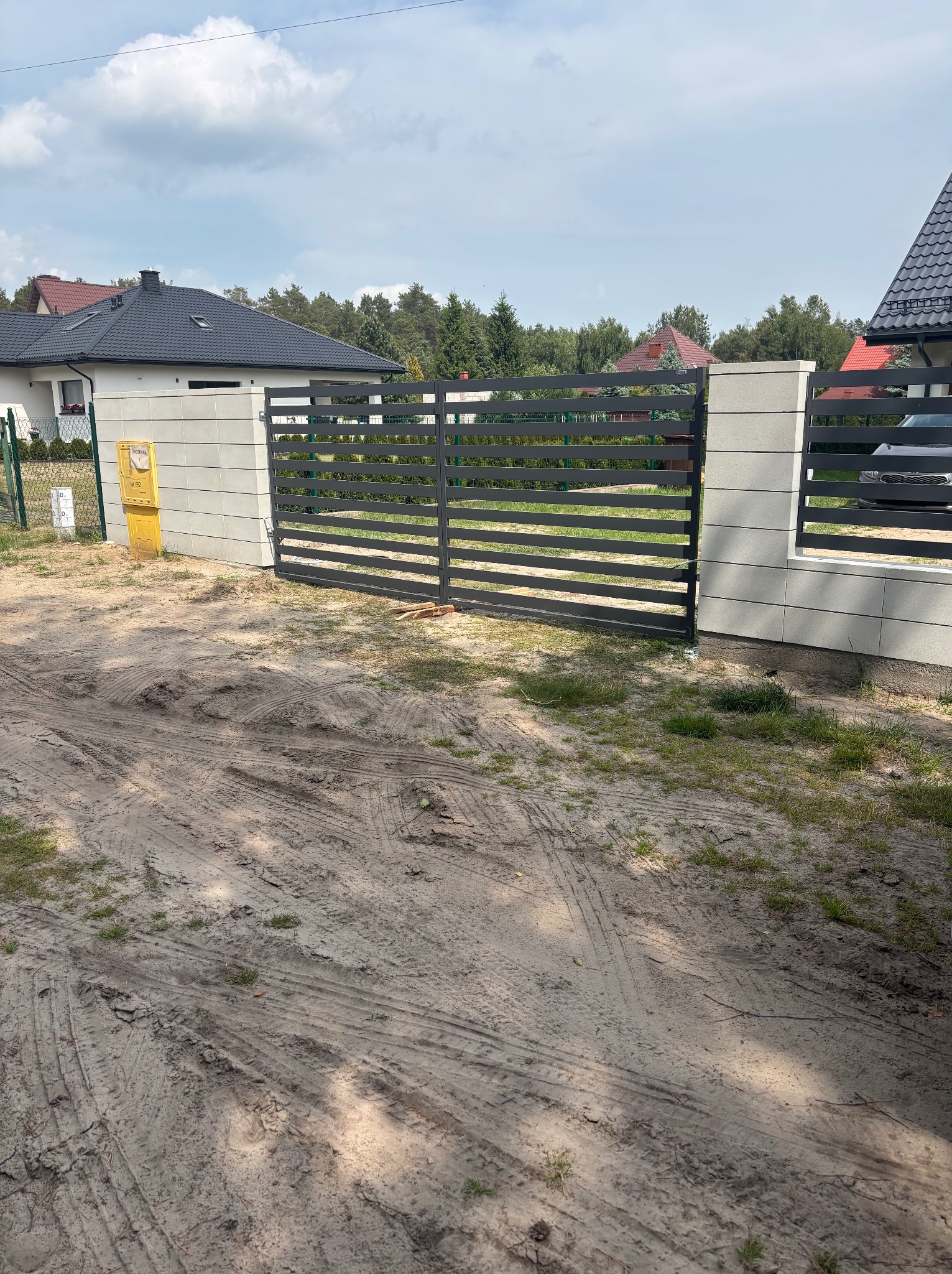 Nowoczesna, pozioma brama wjazdowa z szarych paneli, osadzona w jasnych, betonowych słupkach. Widoczny fragment domu i skrzynka pocztowa.