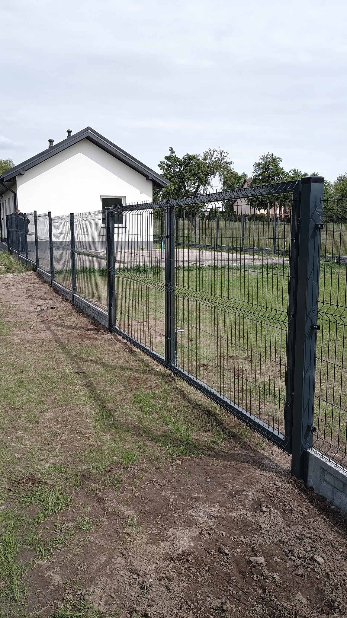 Nowoczesne ogrodzenie panelowe z metalową bramą na tle domu z ogrodem. Widoczne słupki i podmurówka z szarej kostki. Całość w odcieniach szarości i zieleni.