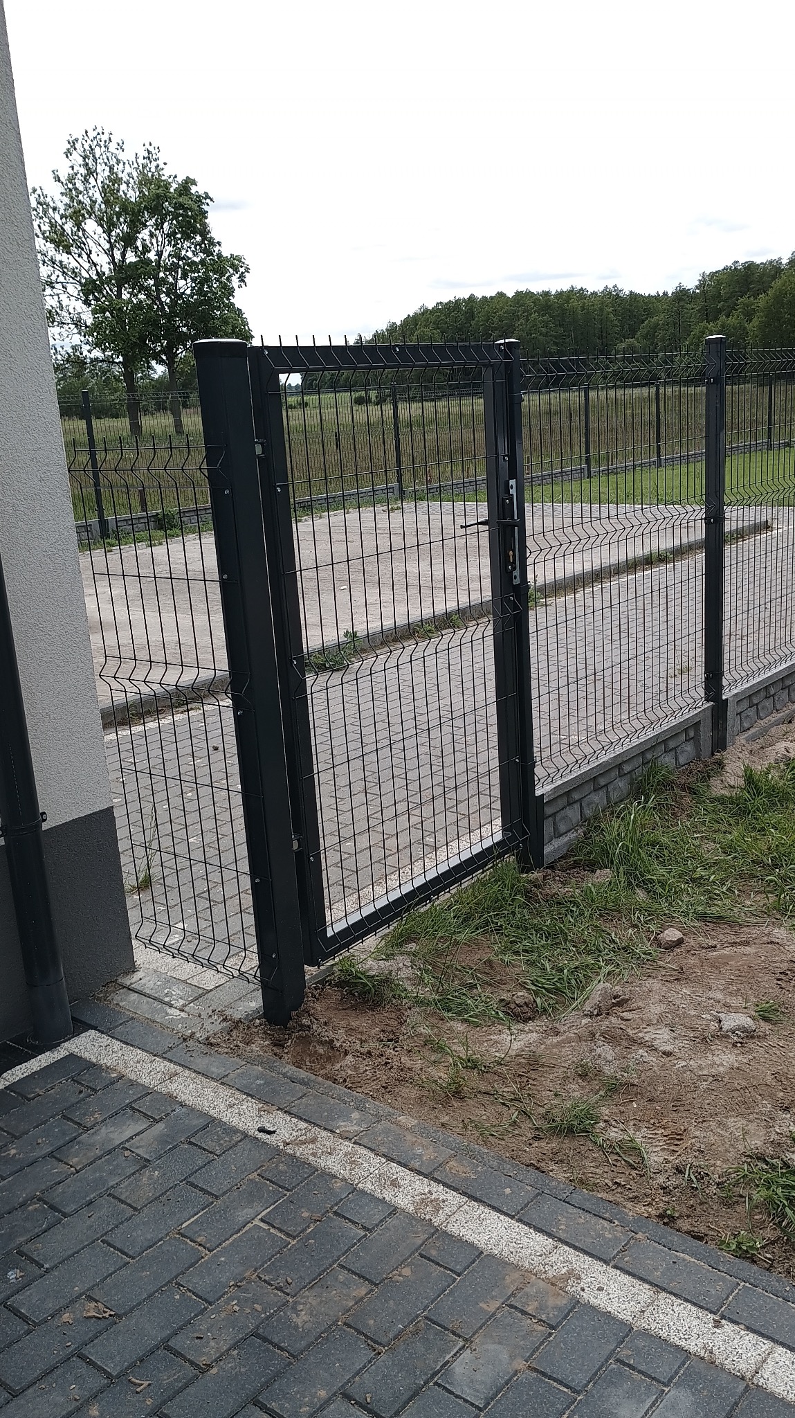 Nowoczesna, metalowa furtka w ogrodzeniu panelowym, z widocznym zamkiem i klamką. Furtka otwarta, prowadzi na ścieżkę z kostki brukowej. Tło: zieleń drzew.