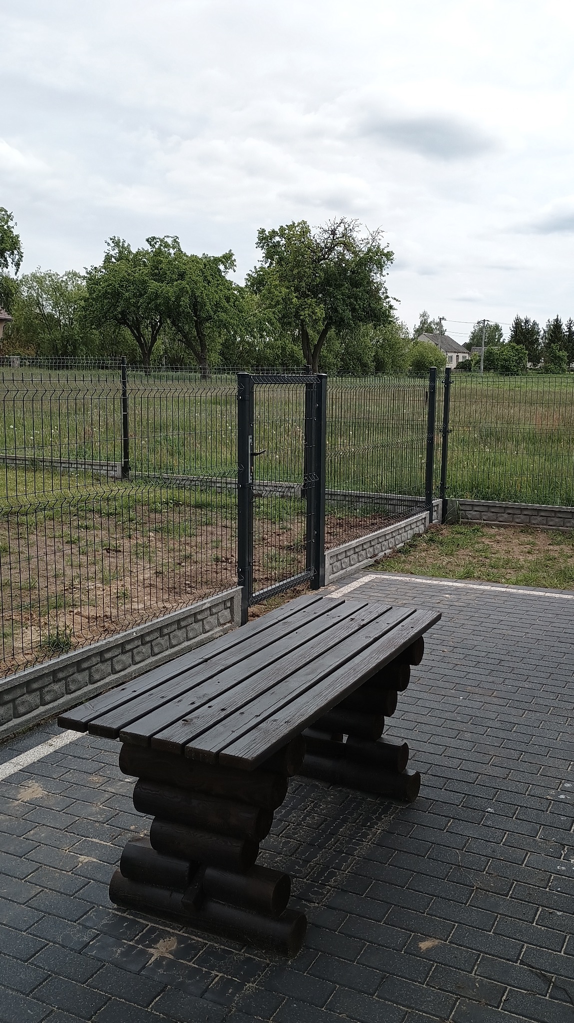 Ciemna ławka z drewnianych bali na kostce brukowej, na tle ogrodzenia panelowego z bramką i zieleni. Pochmurne niebo.