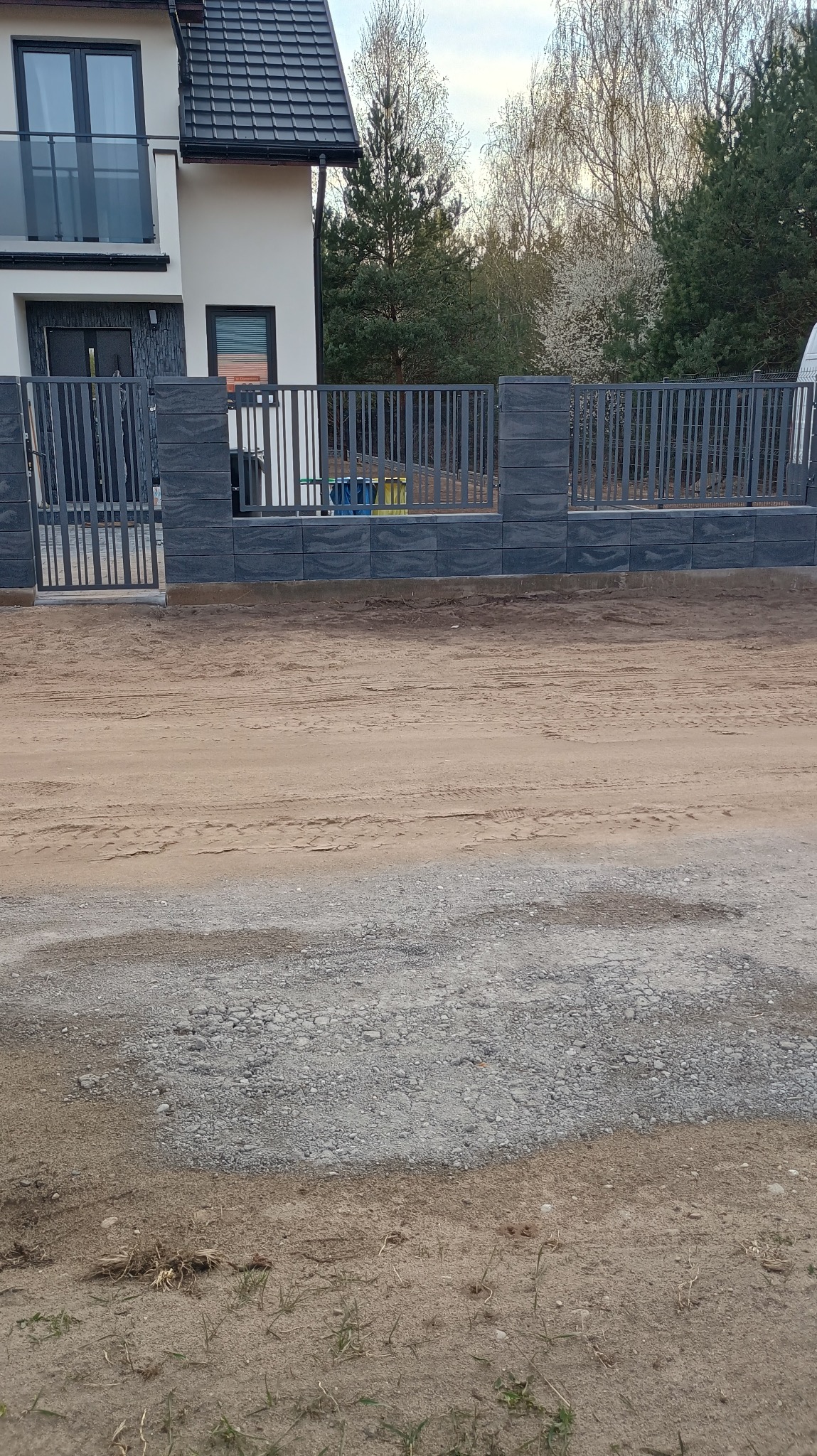 Nowoczesne ogrodzenie panelowe z szarej kostki łupanej i metalowych przęseł na tle domu z czarnym dachem i zieleni. Widoczne ślady opon na piasku.