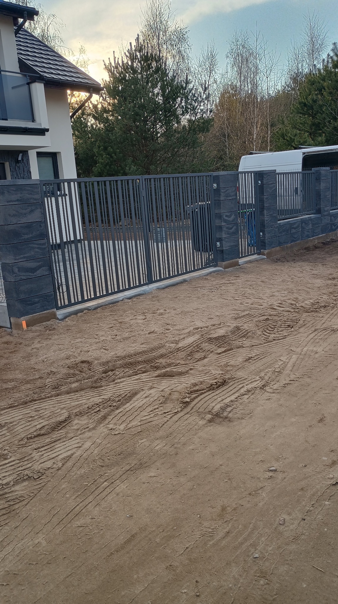 Nowoczesne ogrodzenie panelowe z betonowymi słupkami, widoczne na tle domu i pojazdu dostawczego. Teren wokół ogrodzenia przygotowany pod dalsze prace.