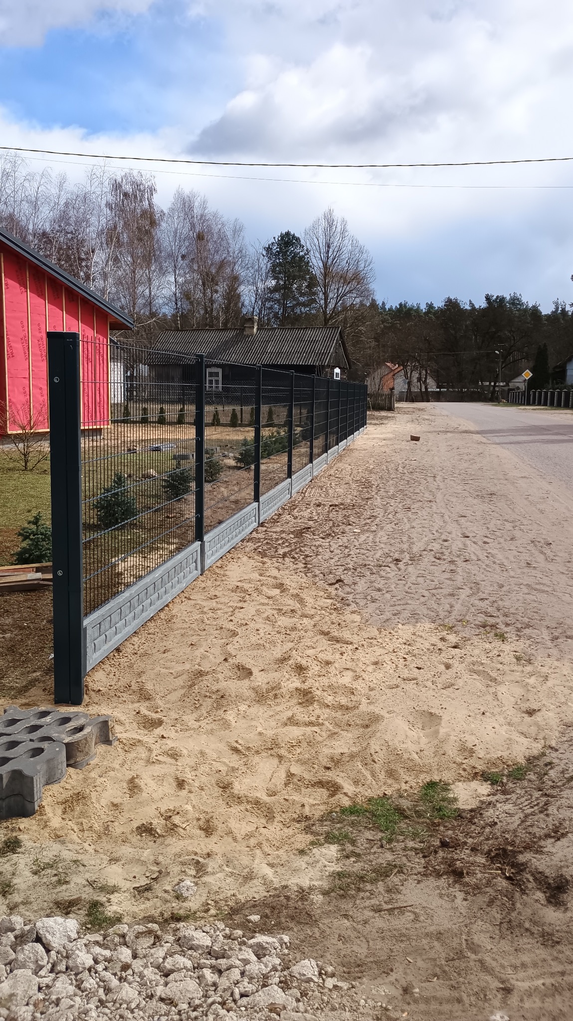 Nowoczesne ogrodzenie panelowe z podmurówką, czarne słupki, widok z boku, piasek na pierwszym planie, dom w tle, błękitne niebo.