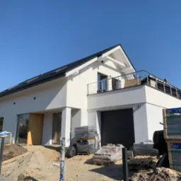 Nowoczesny dom jednorodzinny z płaskim dachem i balkonem, w trakcie prac wykończeniowych; widoczne palety z kostką brukową przed garażem, niezabezpieczone otwory okienne i drzwiowe.