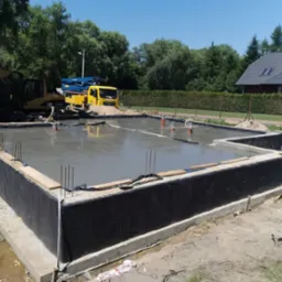 Wylany fundament z izolacją pionową, widoczne wystające pręty zbrojeniowe, pomarańczowe rury, na drugim planie ż&oacute;łty betonow&oacute;z i koparka CAT.