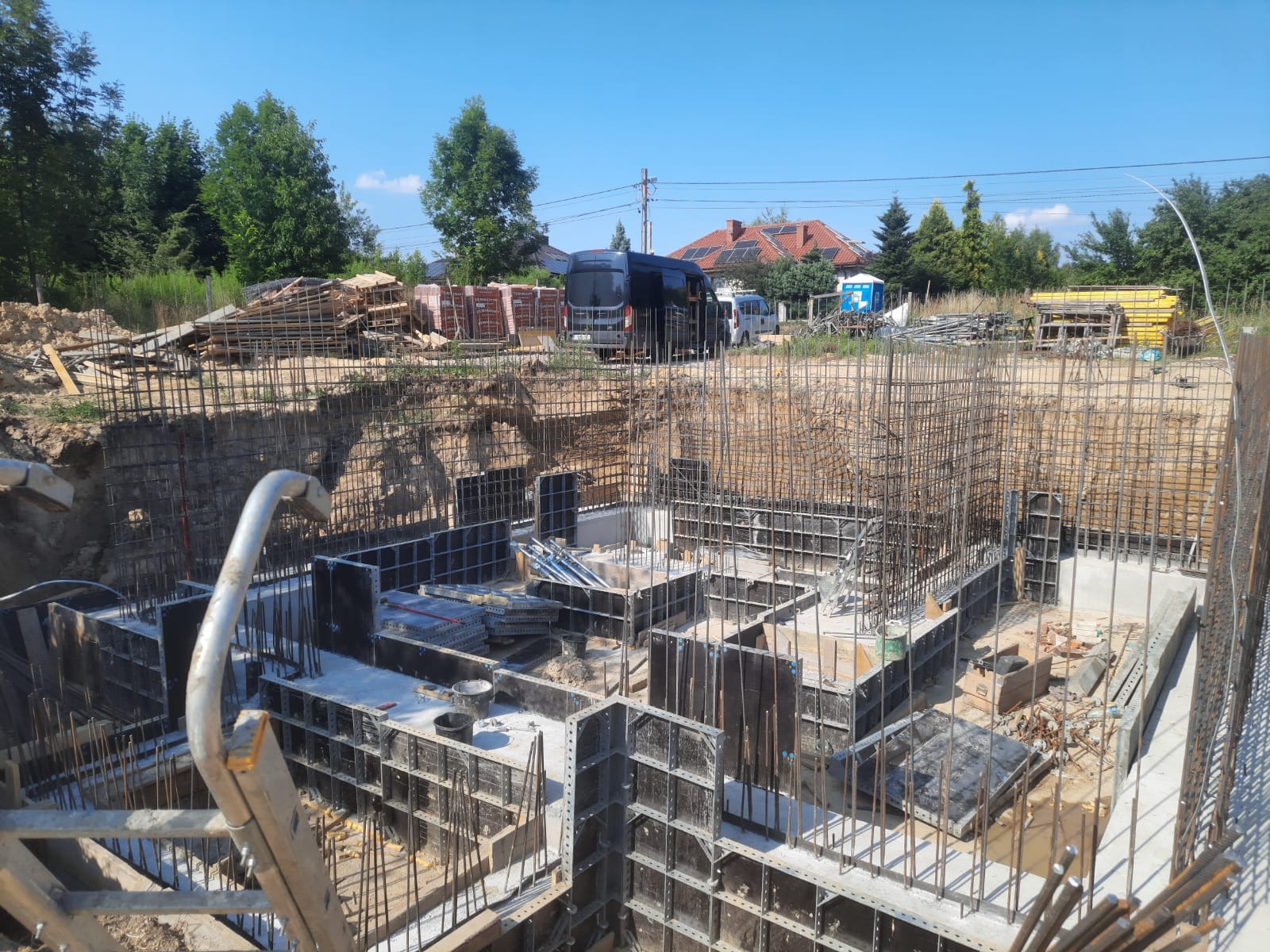 Fundamenty domu w trakcie budowy: szalunki, zbrojenie, częściowo zalany beton, widoczne materiały budowlane i teren budowy.