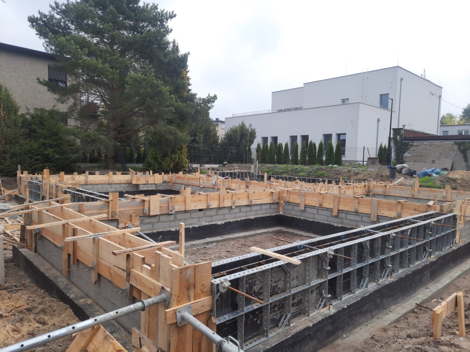 Przygotowanie fundamentów pod budynek: szalunki drewniane i metalowe, izolacja przeciwwilgociowa, bloczki betonowe, widok na plac budowy z budynkami w tle.