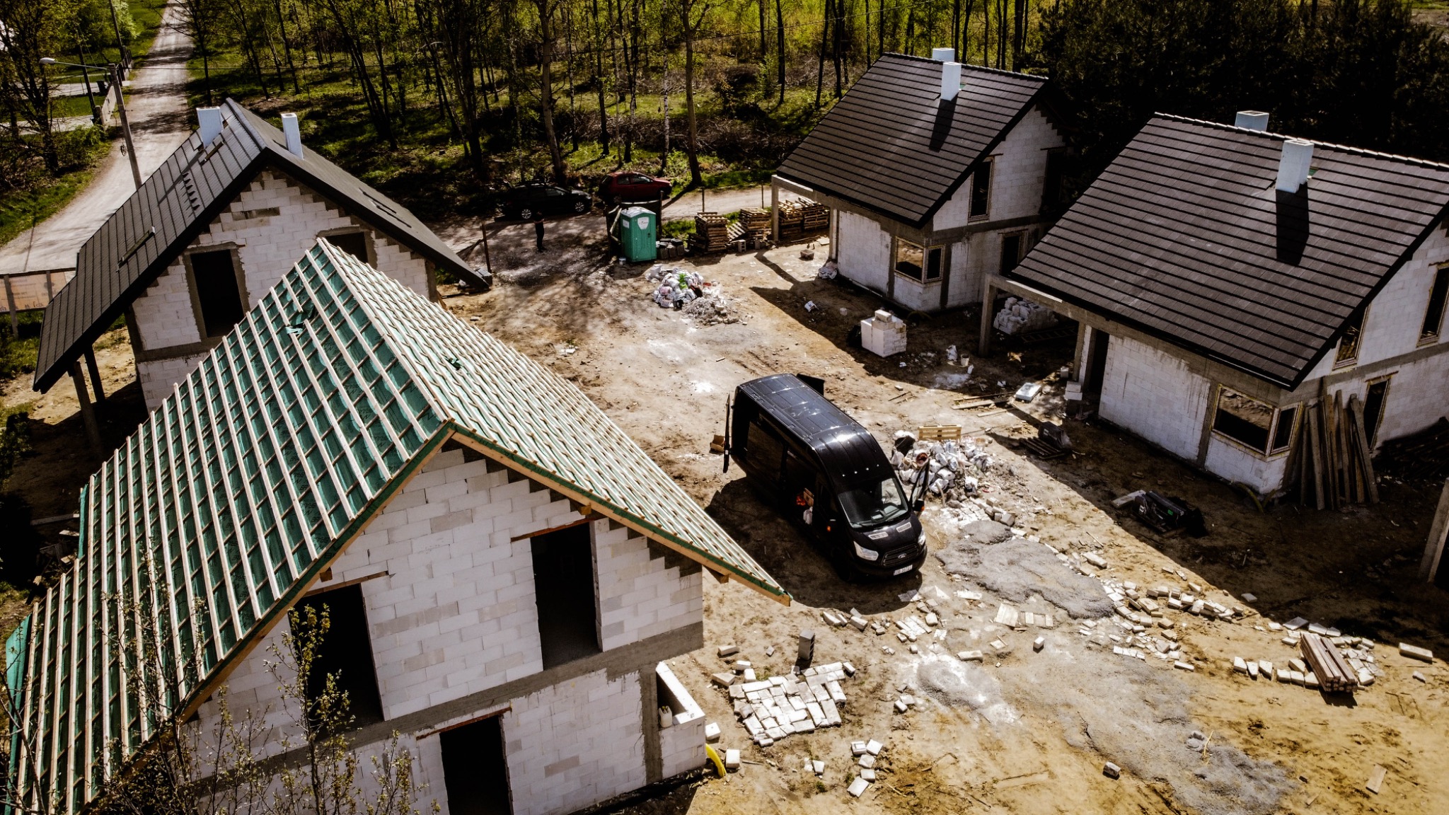 Widok z góry na plac budowy z trzema domami w różnym stopniu zaawansowania: jeden z zielonym szkieletem dachu, drugi w stanie surowym zamkniętym z ciemną dachówką, trzeci z oknami i kominem. Obok...