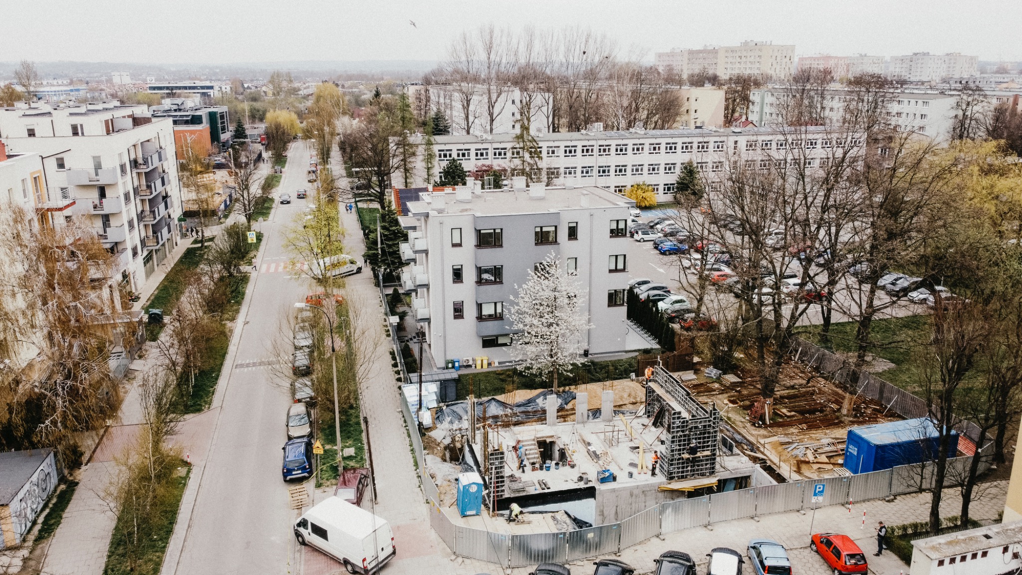 Widok z góry na plac budowy z fundamentami nowego budynku mieszkalnego w otoczeniu istniejącej zabudowy miejskiej, widoczny szalunek i robotnicy w pomarańczowych kamizelkach.