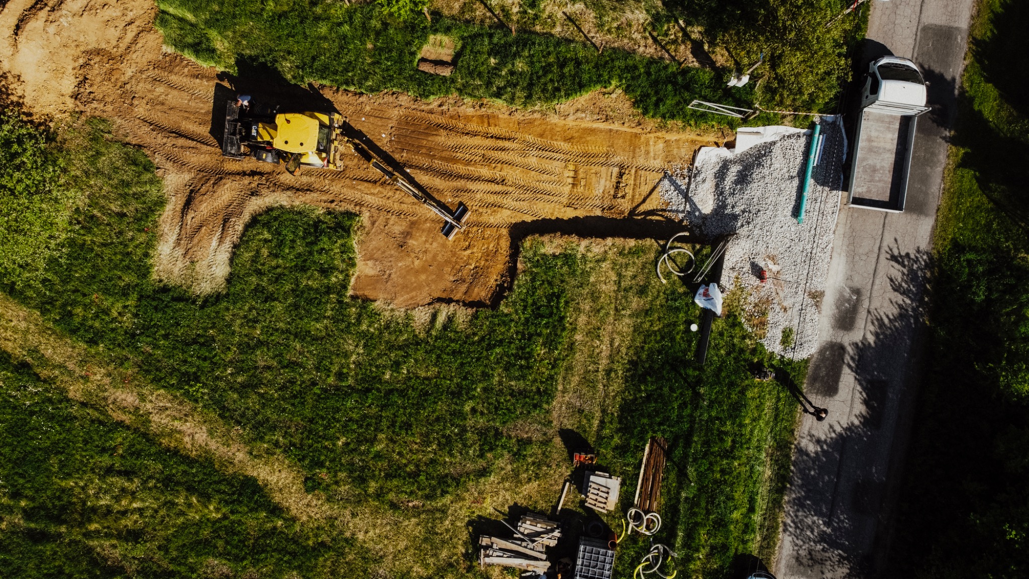 Widok z góry na wykop pod fundamenty, z żółtą koparką w trakcie pracy, obok składowany żwir i podjeżdżający samochód ciężarowy, otoczone trawnikiem i drzewami.