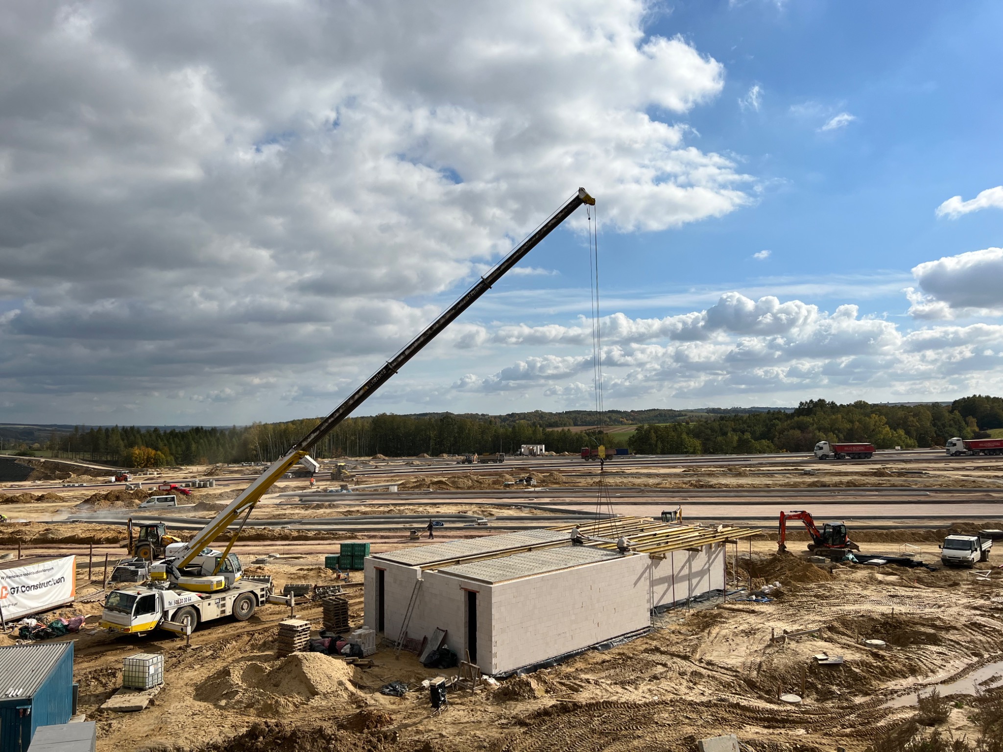 Budowa modułowego domu z betonu komórkowego: dźwig podnosi element dachu na budynek, widoczne ciężarówki w tle, krajobraz zalesiony, plac budowy w trakcie prac.