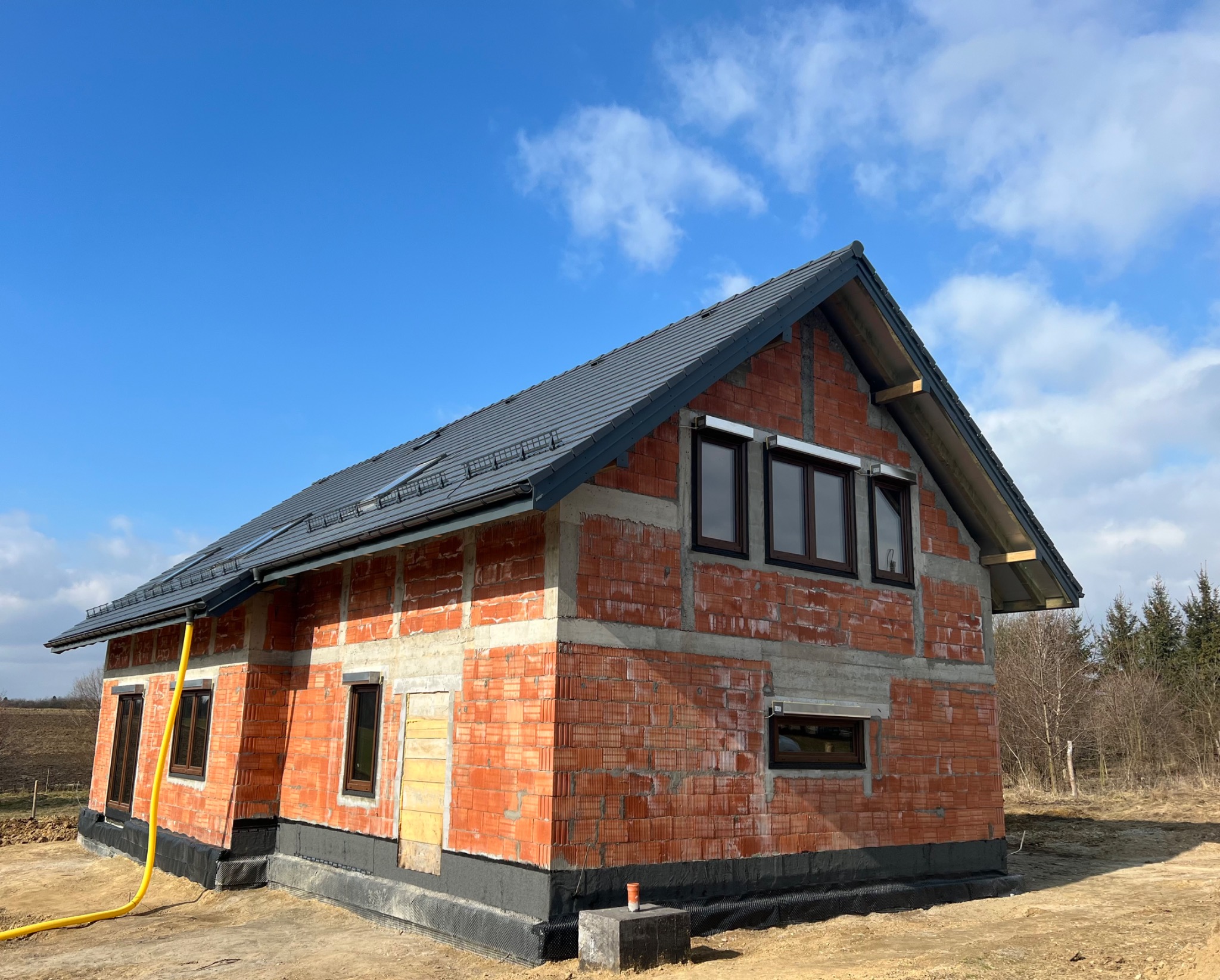 Surowy stan domu z czerwonej cegły z szarą dachówką na tle błękitnego nieba z chmurami, widoczne fundamenty z izolacją, żółta rura spustowa i zamurowane okno.