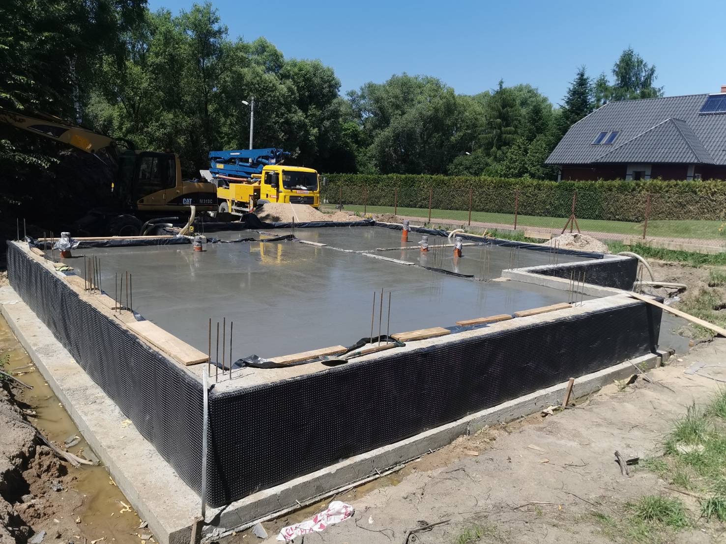 Wylany fundament z izolacją pionową, widoczne wystające pręty zbrojeniowe, pomarańczowe rury, na drugim planie żółty betonowóz i koparka CAT.