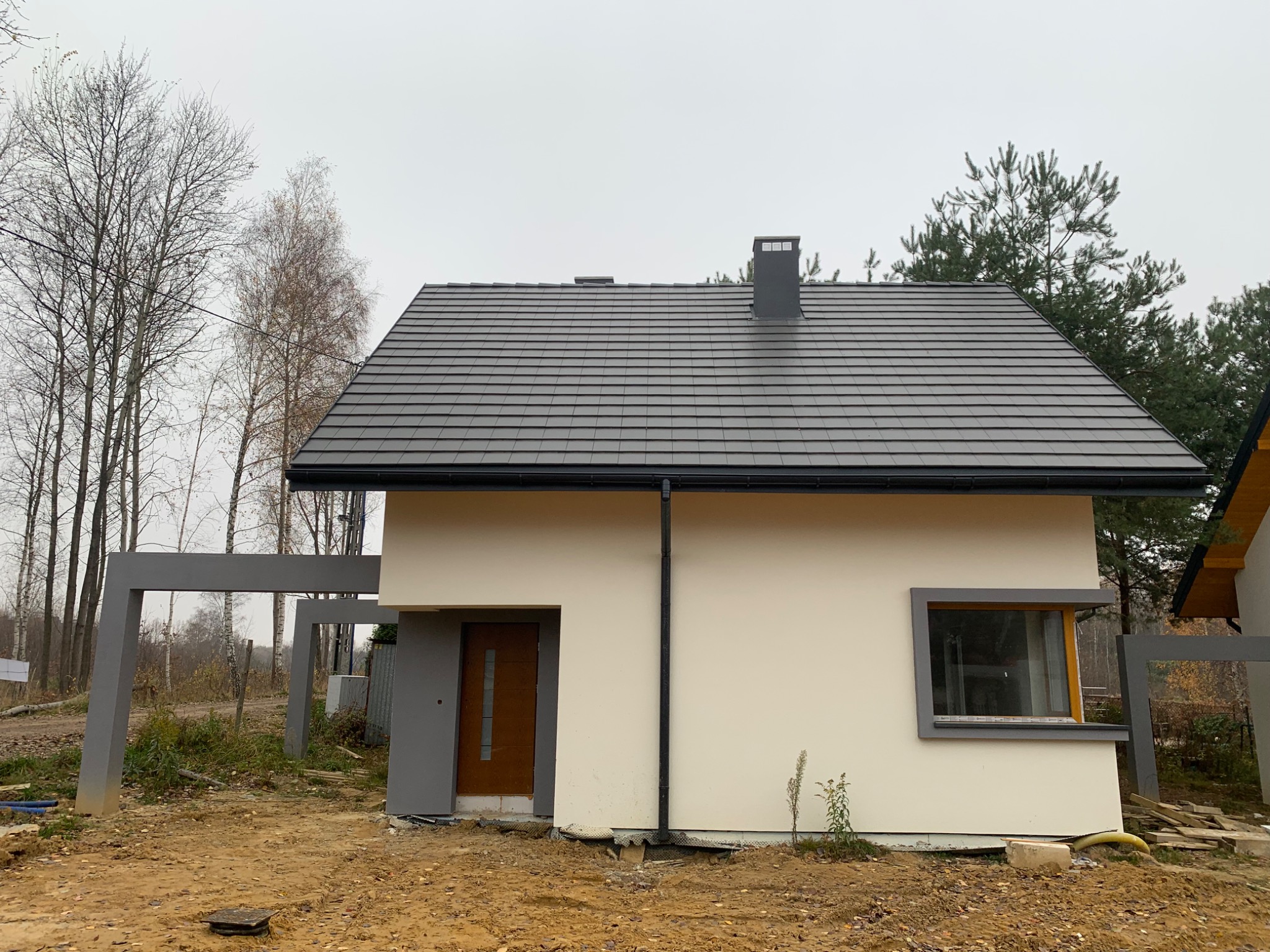 Niedawno wybudowany, parterowy dom z ciemną dachówką, szarą stolarką okienną i brązowymi drzwiami wejściowymi, na działce w stanie surowym, w tle widoczne drzewa bez liści.