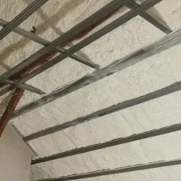 Poddasze z widoczną konstrukcją metalową i wypełnieniem z piany izolacyjnej, widoczne fragmenty ściany i żółtej folii ochronnej.