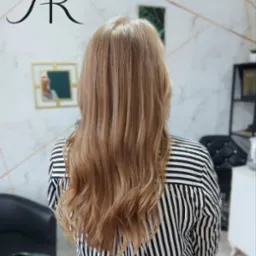 Tył głowy kobiety z długimi, falowanymi włosami w odcieniach blondu, ubrana w bluzkę w czarno-białe paski, widok na salon fryzjerski w tle.