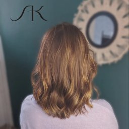 AK Studio Włosów - Tył głowy kobiety z brązowymi, pofalowanymi włosami z blond refleksami, na tle ściany w kolorze butelkowej zieleni i okrągłego lustra z dekoracyjną ramą.