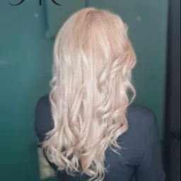 Tył głowy kobiety z długimi, blond, pofalowanymi włosami, na tle ciemnozielonej ściany z widocznym logo firmy w lewym górnym rogu.