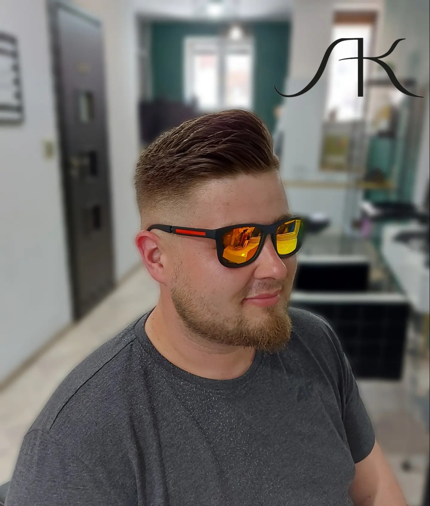 Mężczyzna z modną fryzurą typu fade i stylizowaną górą, noszący okulary przeciwsłoneczne z pomarańczowymi szkłami, w tle salon z logo.