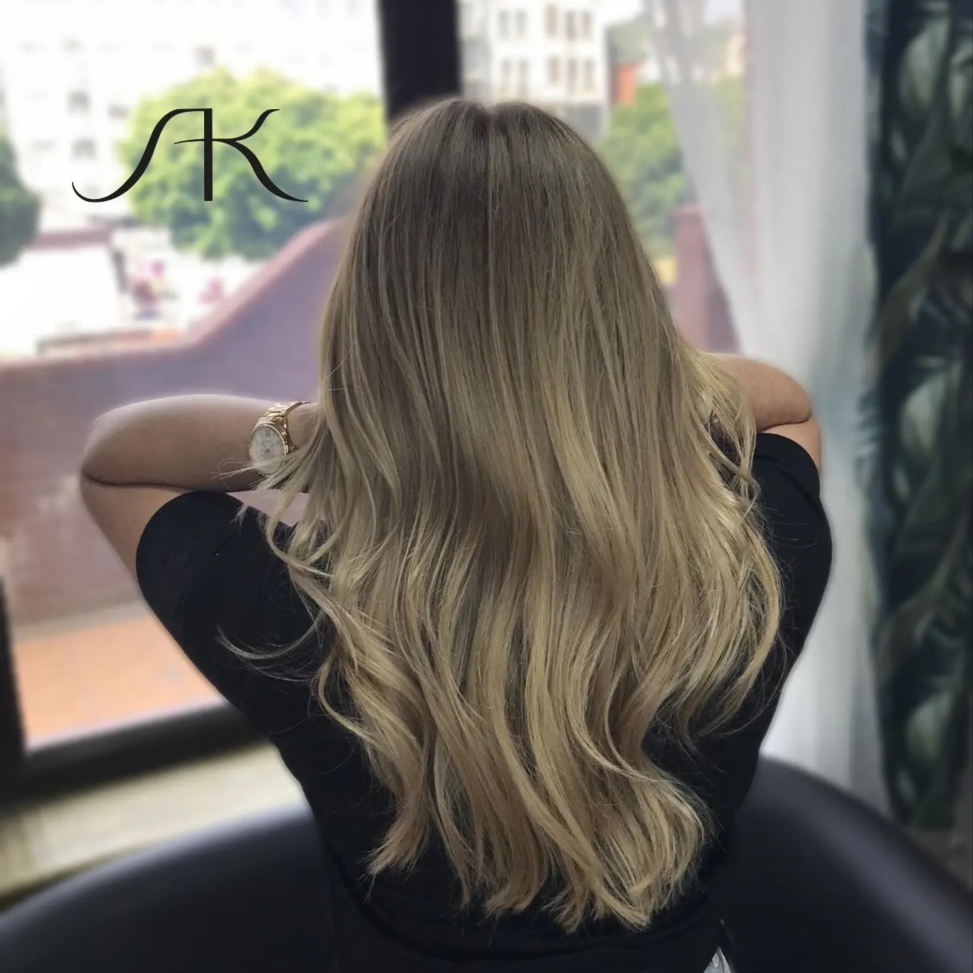 Długie, falowane włosy w odcieniach blondu, widok z tyłu, z subtelnym logo w rogu i widokiem na okno w tle.