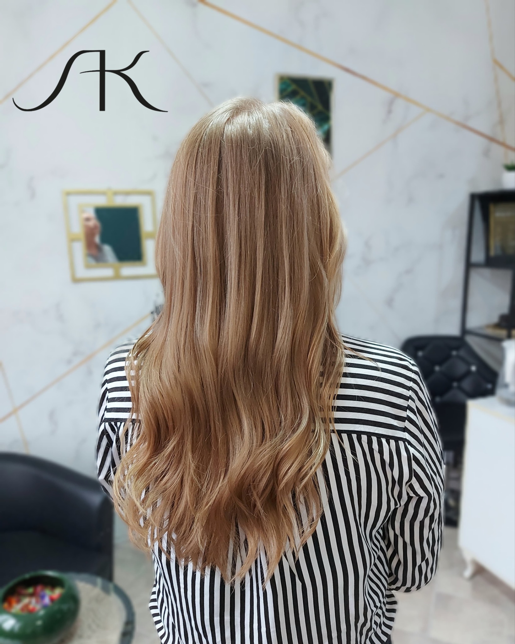 Tył głowy kobiety z długimi, falowanymi włosami w odcieniach blondu, ubrana w bluzkę w czarno-białe paski, widok na salon fryzjerski w tle.