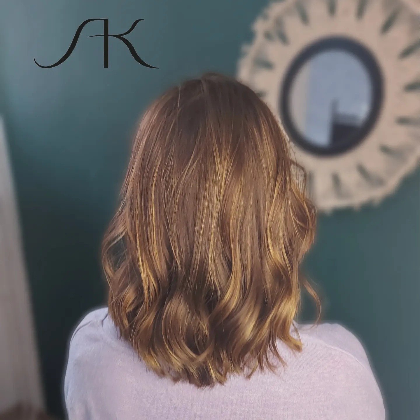 Tył głowy kobiety z brązowymi, pofalowanymi włosami z blond refleksami, na tle ściany w kolorze butelkowej zieleni i okrągłego lustra z dekoracyjną ramą.