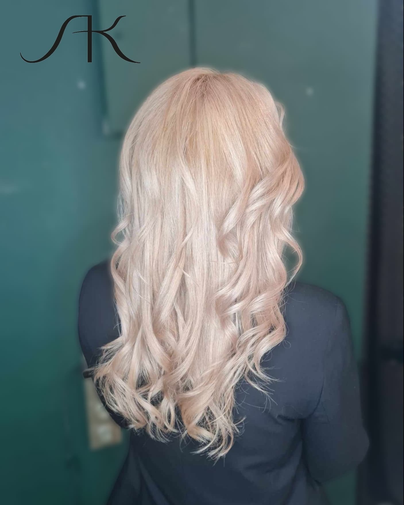 Tył głowy kobiety z długimi, blond, pofalowanymi włosami, na tle ciemnozielonej ściany z widocznym logo firmy w lewym górnym rogu.