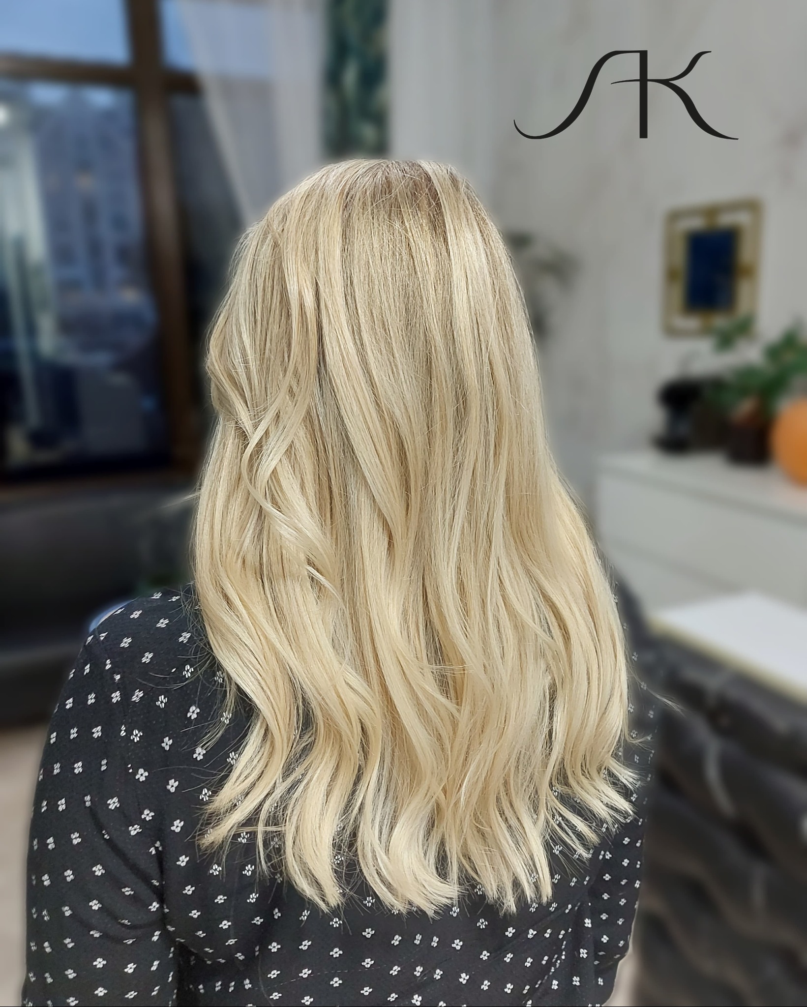 Tył głowy kobiety z długimi, blond, falowanymi włosami po koloryzacji i stylizacji, widoczne logo salonu.