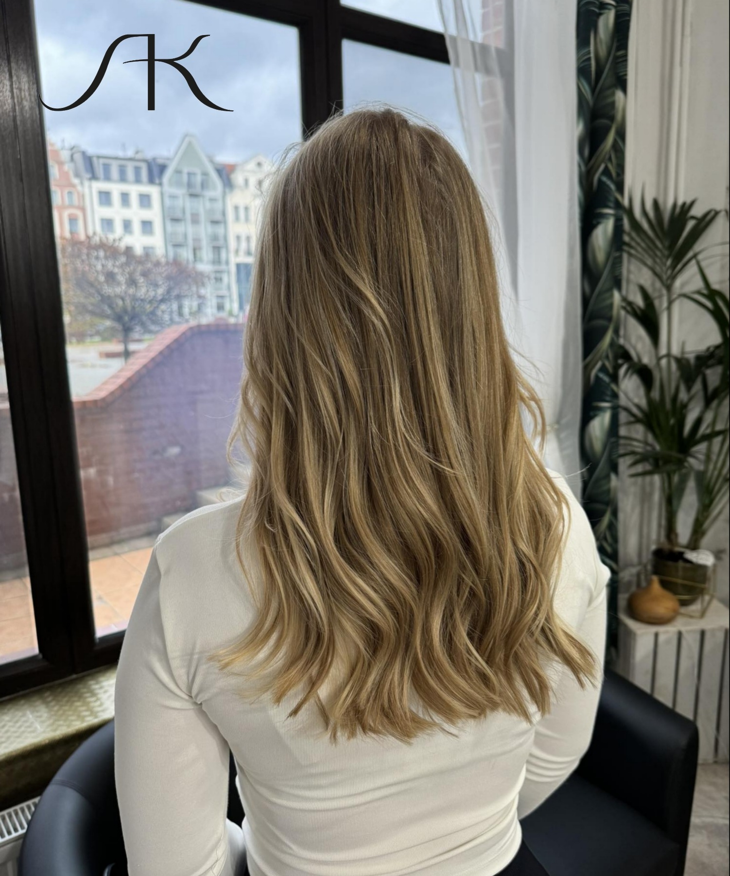 Długie, blond włosy z delikatnymi falami, widok od tyłu, na tle okna z widokiem na miasto i rośliny doniczkowe.