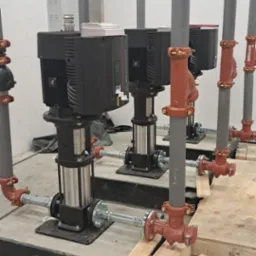 Rząd pionowych pomp przemysłowych, czarne silniki i srebrne korpusy, połączone rurami z czerwonymi zaworami w surowym, betonowym pomieszczeniu.