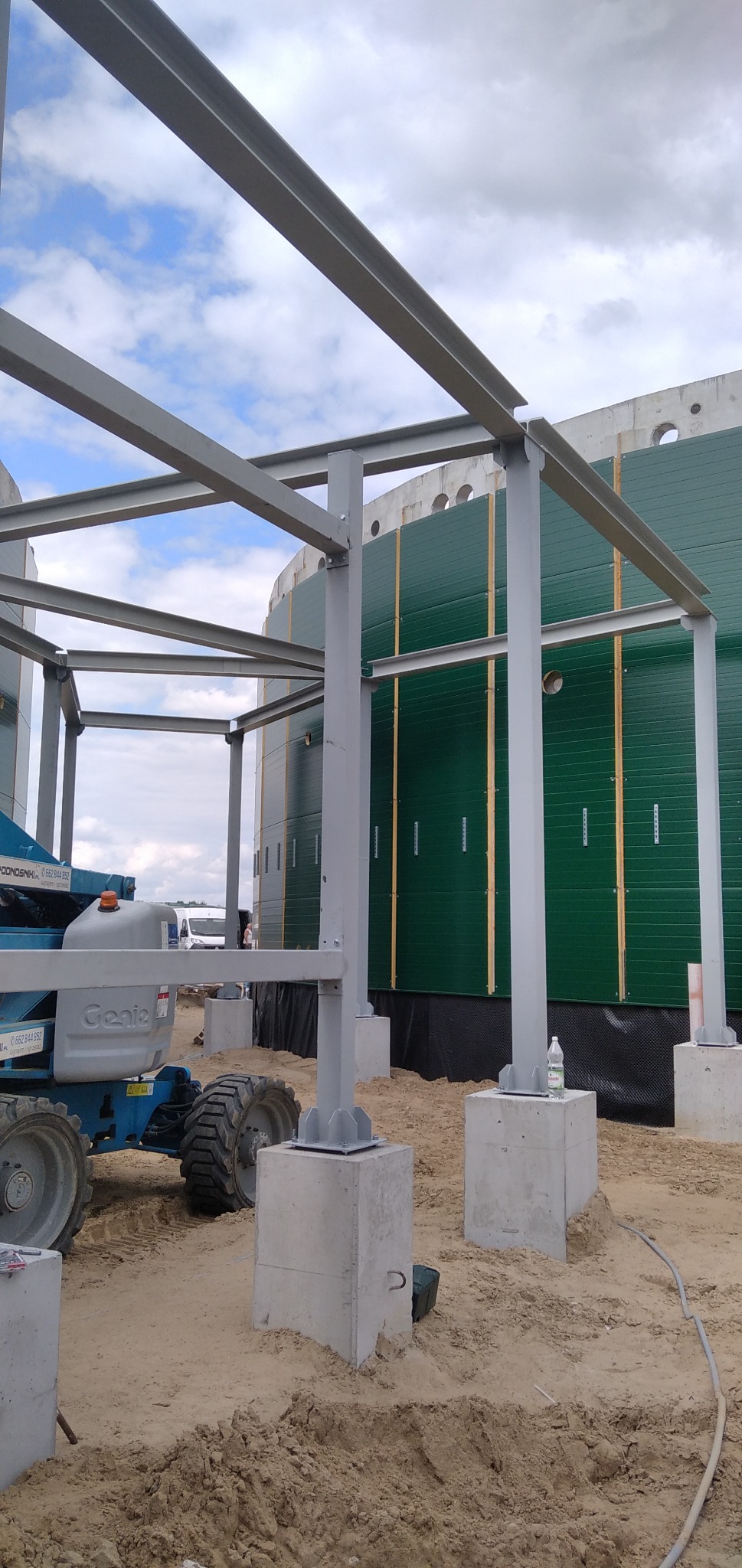 Montaż stalowej konstrukcji szkieletowej hali, widoczny podnośnik koszowy, betonowe fundamenty i fragment ściany z zielonej blachy trapezowej.