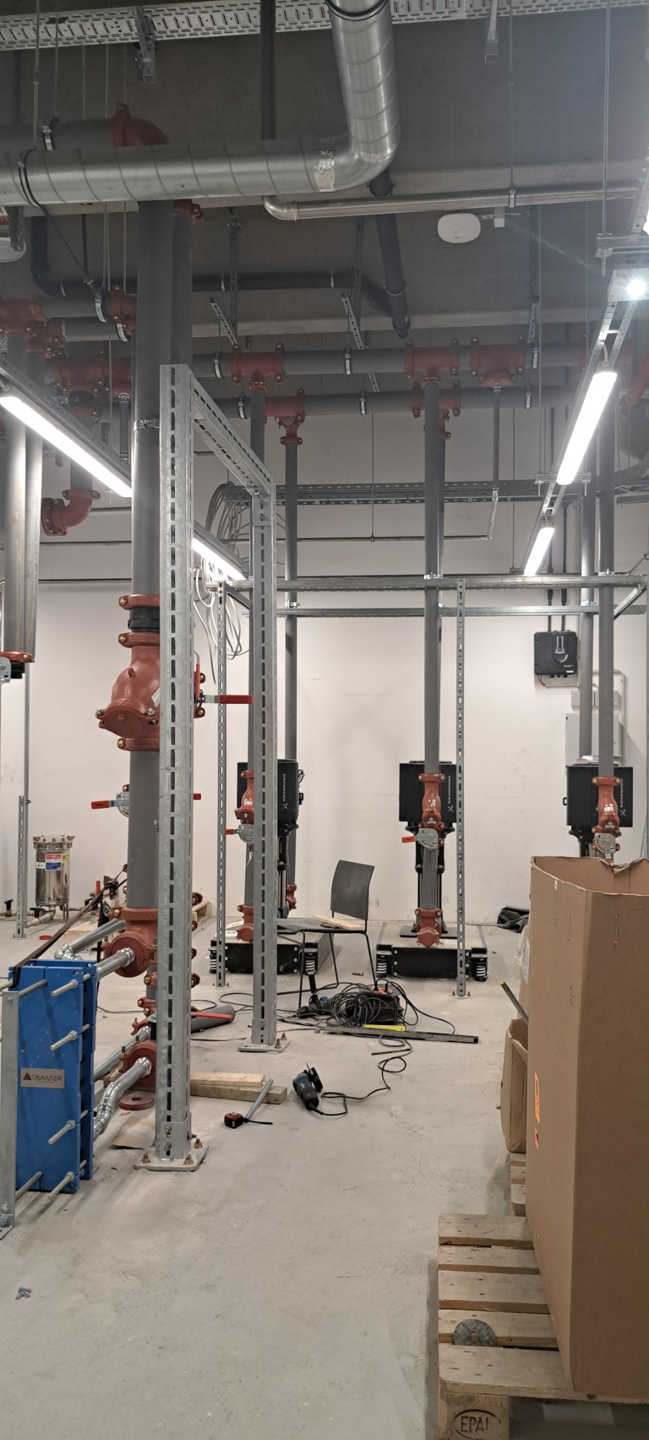 Instalacja przemysłowa w trakcie montażu: rury, zawory z czerwonymi pokrętłami, metalowe konstrukcje wsporcze, narzędzia, kartonowe pudło i krzesło na tle białej ściany i betonowej podłogi.