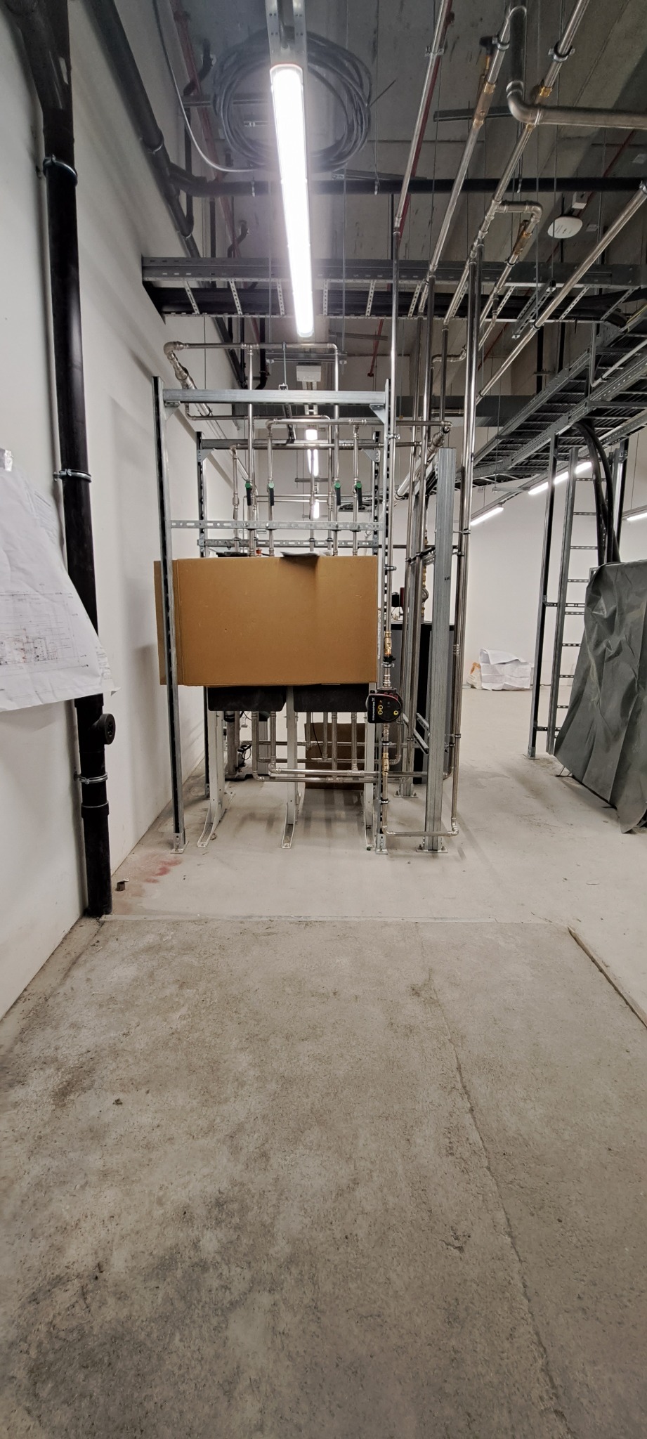 Instalacja rur i armatury w surowym stanie, widoczne rury stalowe, zawory, pompy i osprzęt, częściowo zakryte kartonem, na tle betonowej posadzki i białych ścian w trakcie prac wykończeniowych.