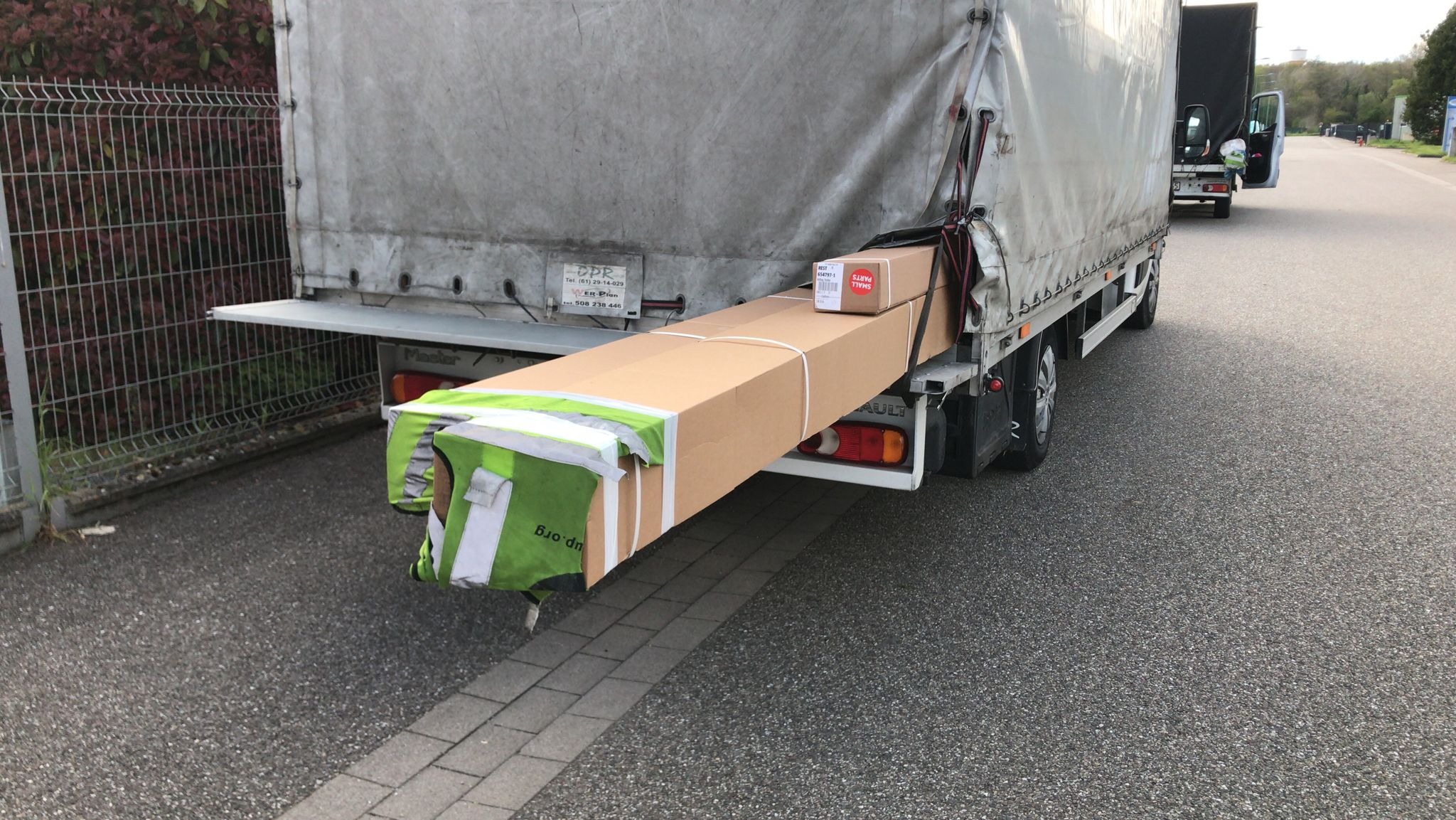 Tył samochodu dostawczego z plandeką, na platformie załadunkowej zabezpieczone pasami znajdują się kartonowe paczki, w tym jedna oznaczona jako 'SMALL PARTS'