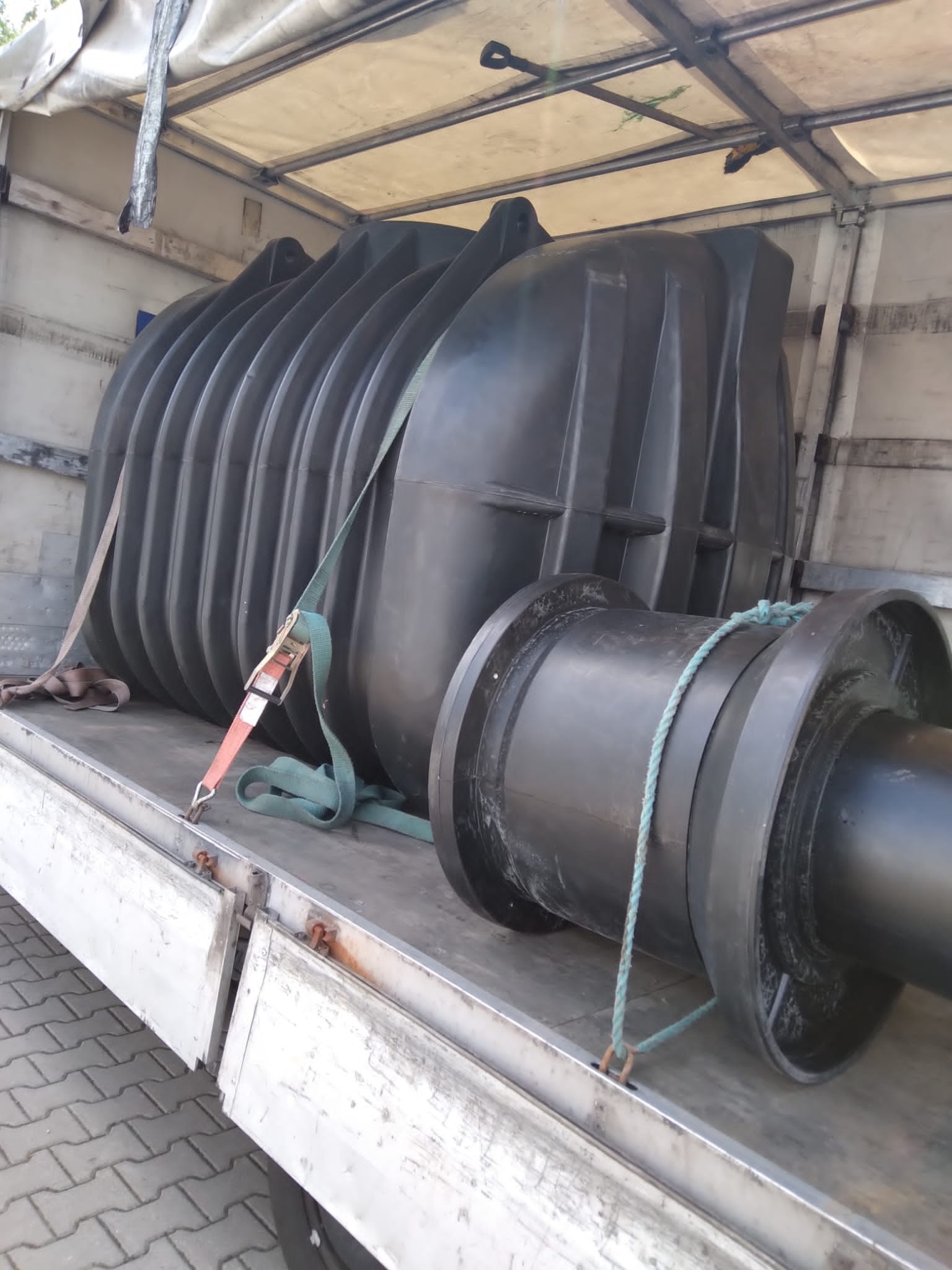 Duży, czarny zbiornik z tworzywa sztucznego, zabezpieczony pasami, transportowany w przestrzeni ładunkowej pojazdu z plandeką.