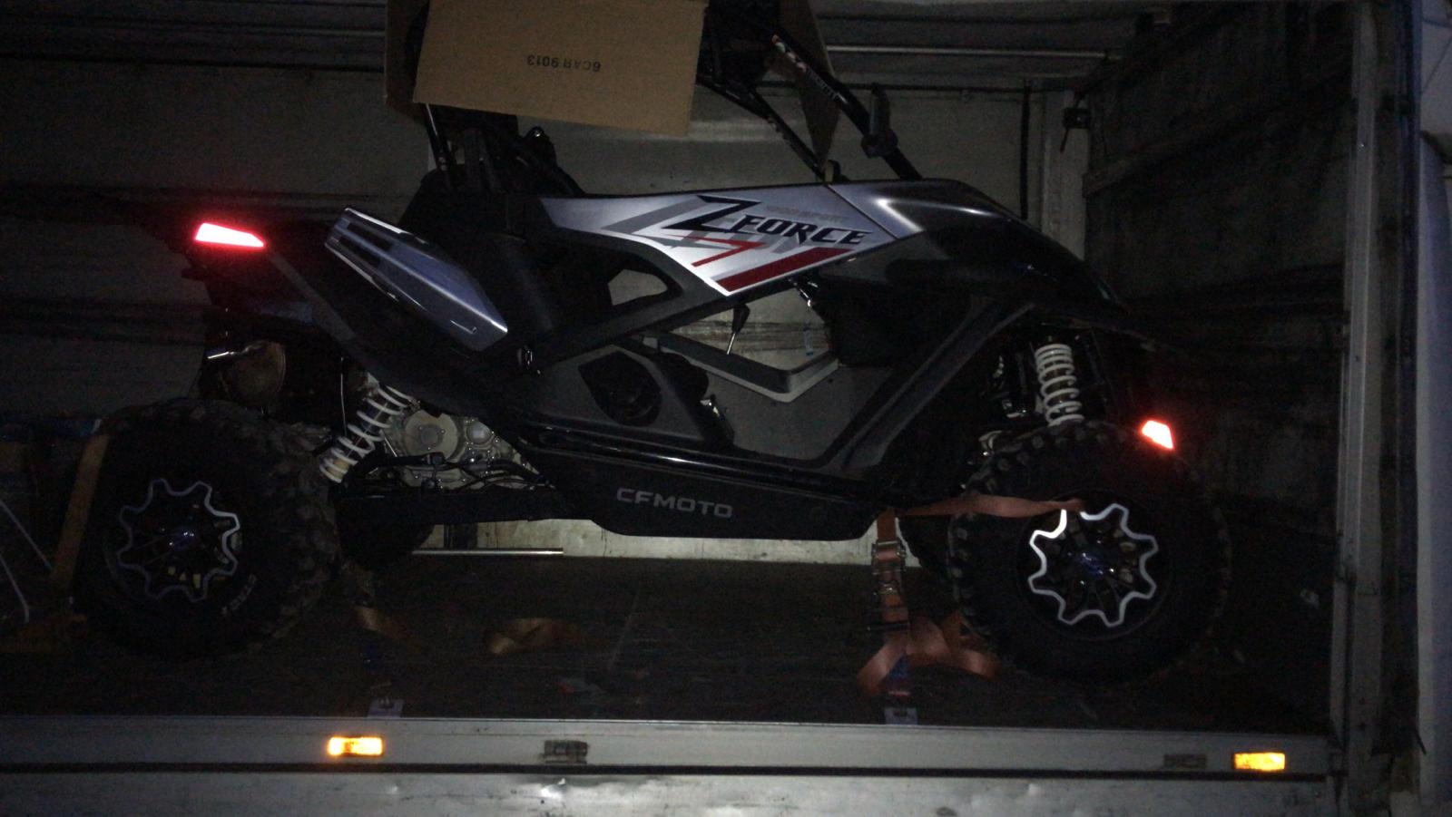 Czarny pojazd ATV marki CFMOTO ZFORCE zabezpieczony pomarańczowymi pasami transportowymi wewnątrz ciemnej przestrzeni ładunkowej busa.